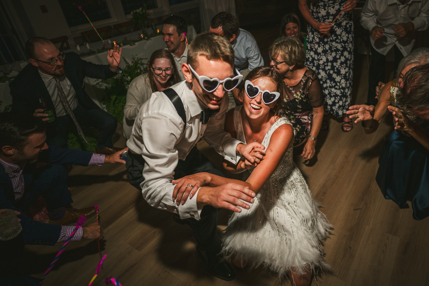 People dancing and celebrating indoors, two central figures wearing sunglasses and smiling, surrounded by others enjoying the event.