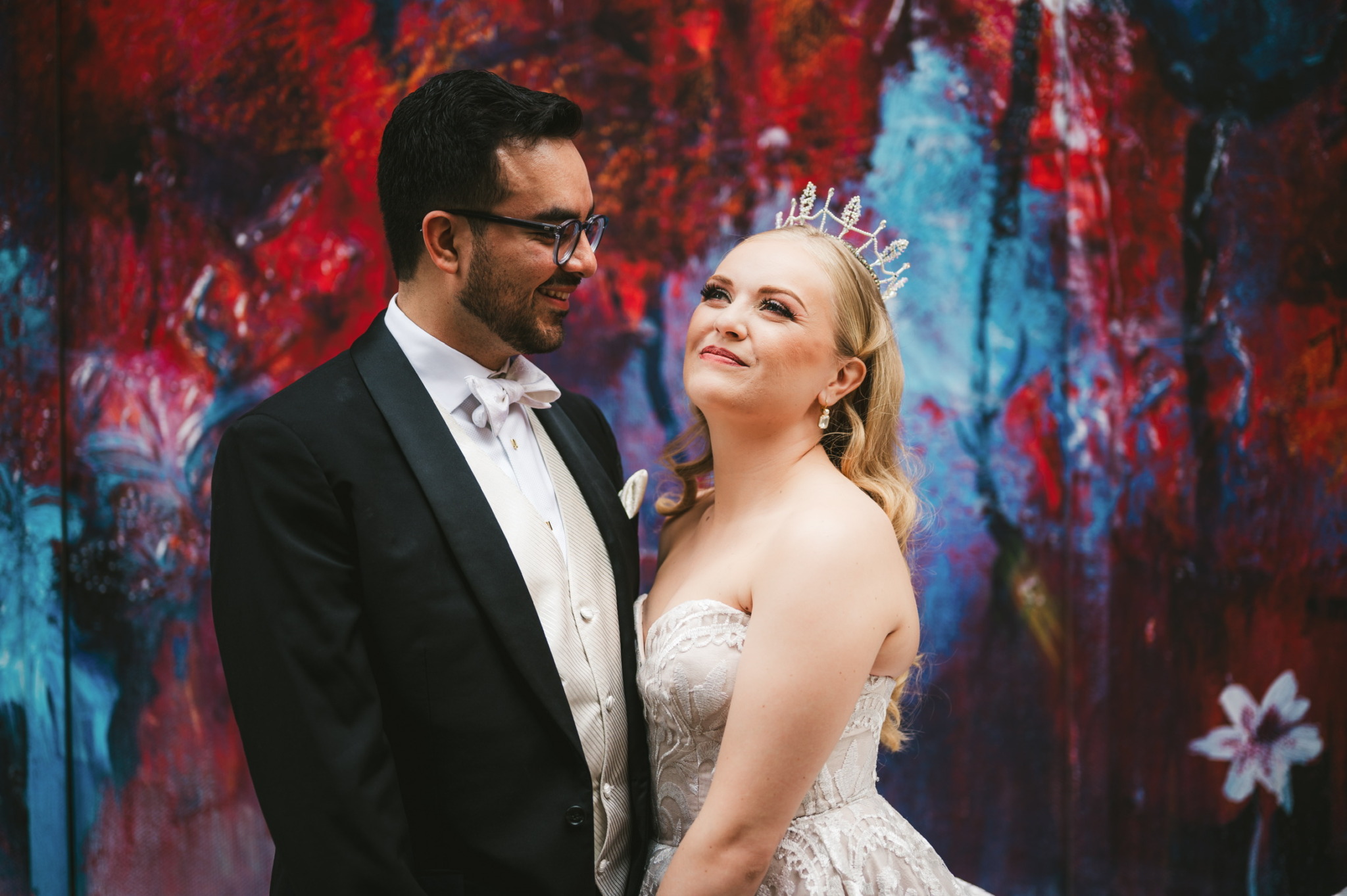 A bride and groom stand close together, smiling, with a colorful abstract background behind them.
