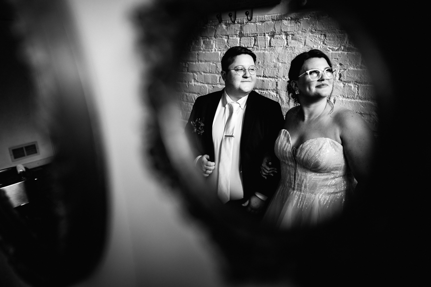 Two people, a man and a woman, stand against a brick wall, looking to the right, in black and white.