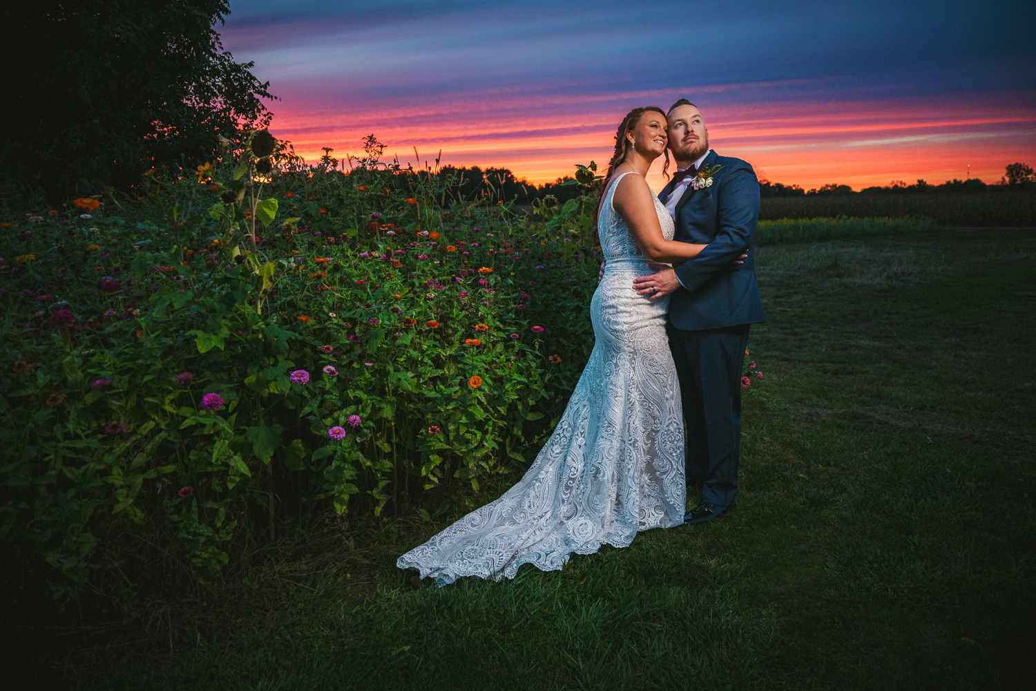 Bride and groom standing outdoors during sunset, embracing each other near a flower bush and open field.