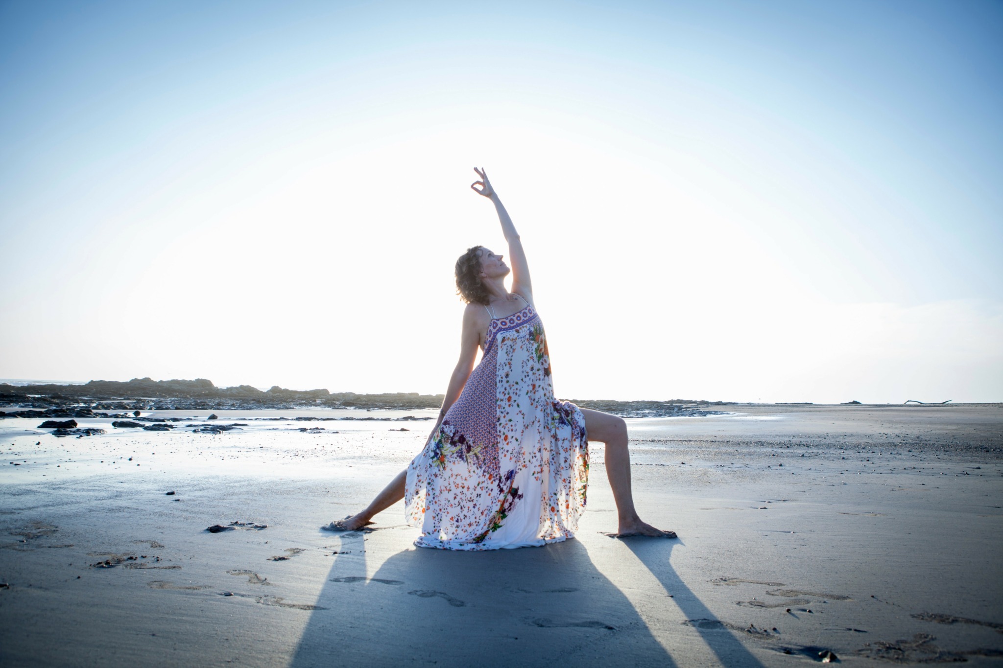 Person sitting on sand with one arm raised, wearing a flowing dress, at a beach during sunset or sunrise.
