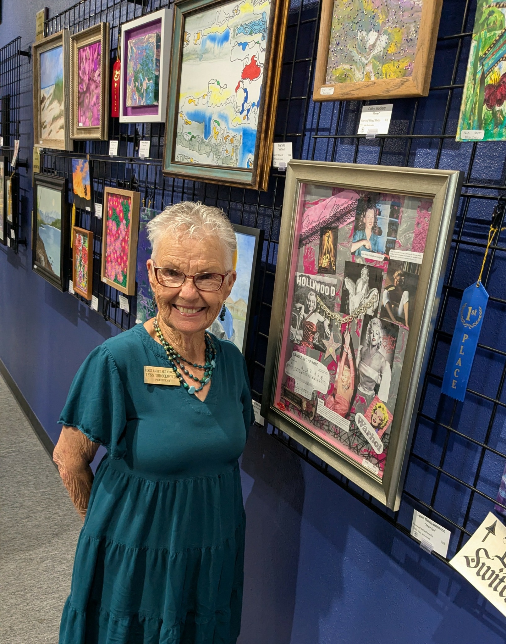 Smiling elderly woman with short gray hair and glasses, standing in front of colorful framed artwork at an art exhibit.