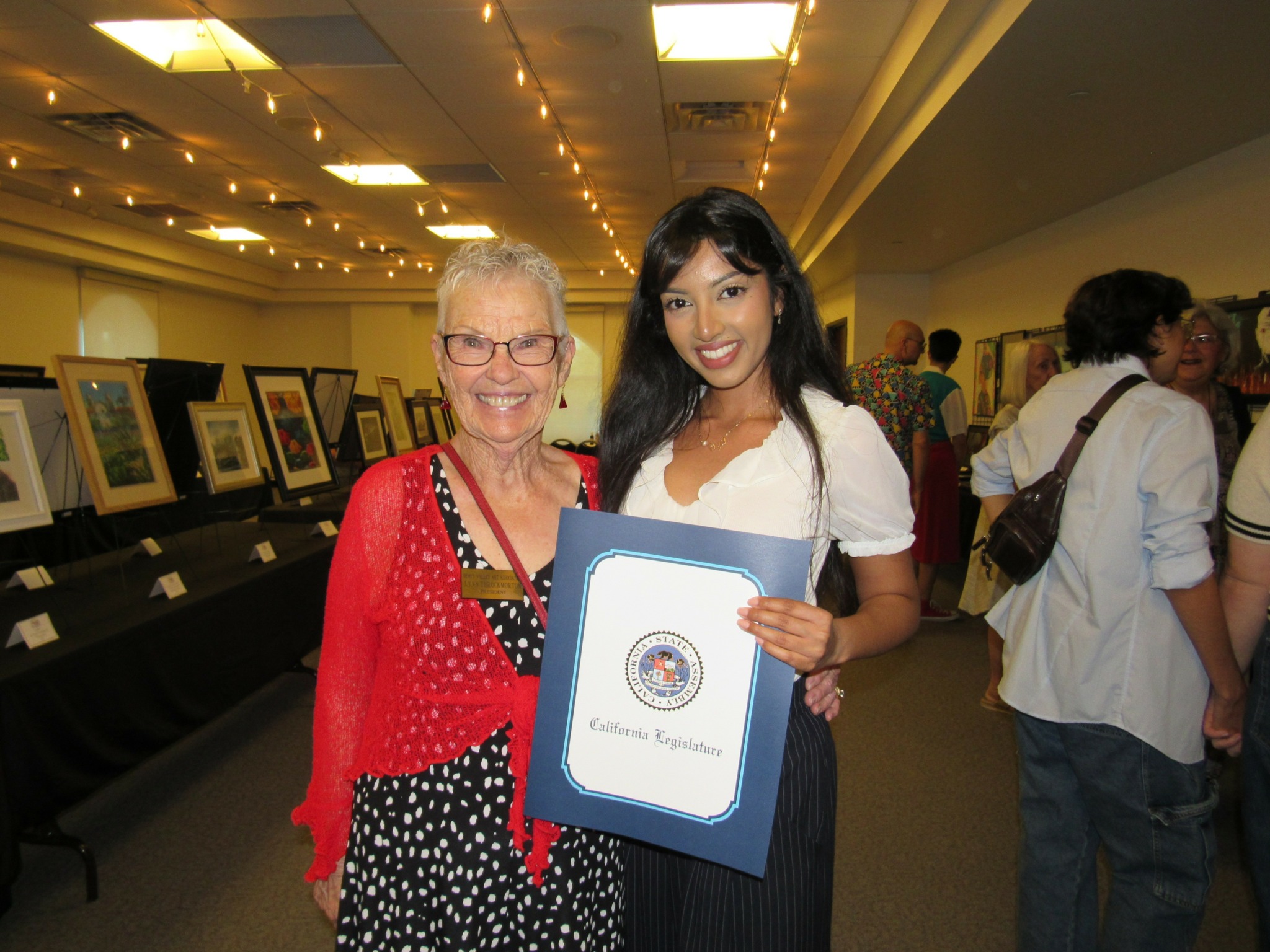 Two women smiling, one holding a certificate, in an indoor gallery with framed artwork displayed on tables.