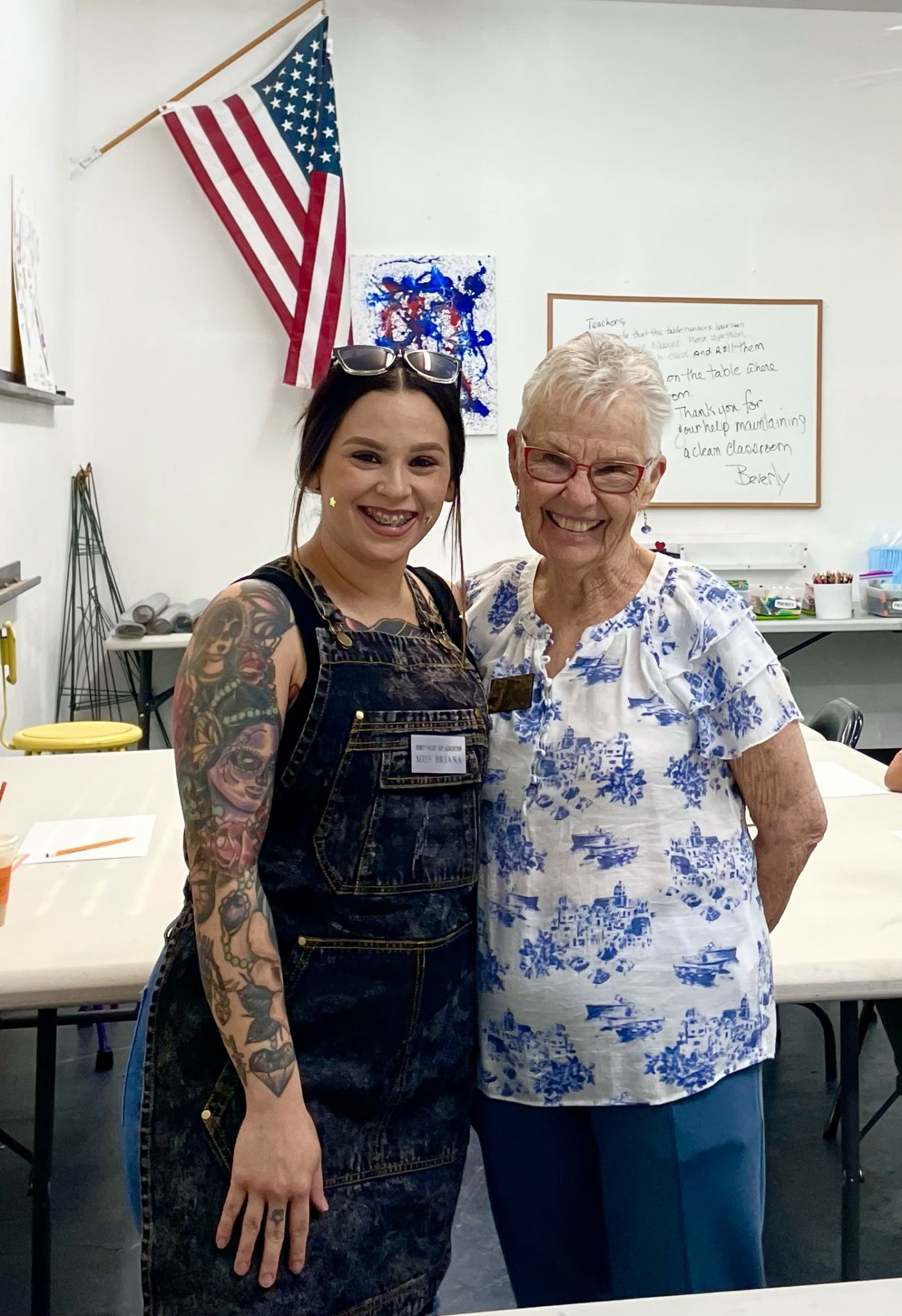 Two women standing together in a room with American flag and whiteboard in background, smiling at the camera.