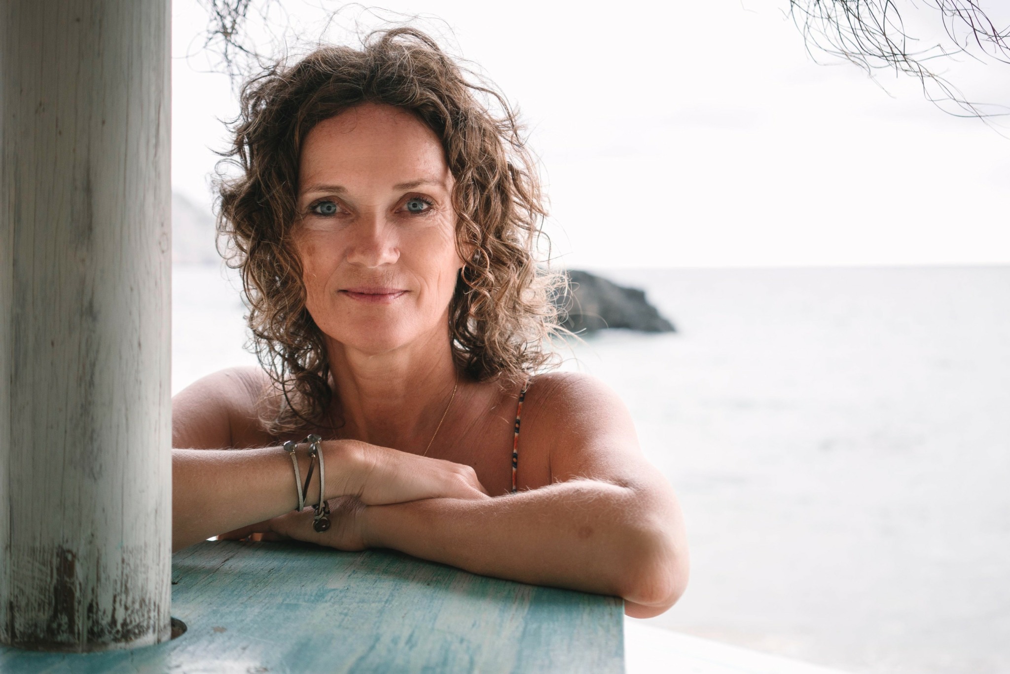 Woman with curly hair leaning on a wooden surface near a beach, smiling at the camera.