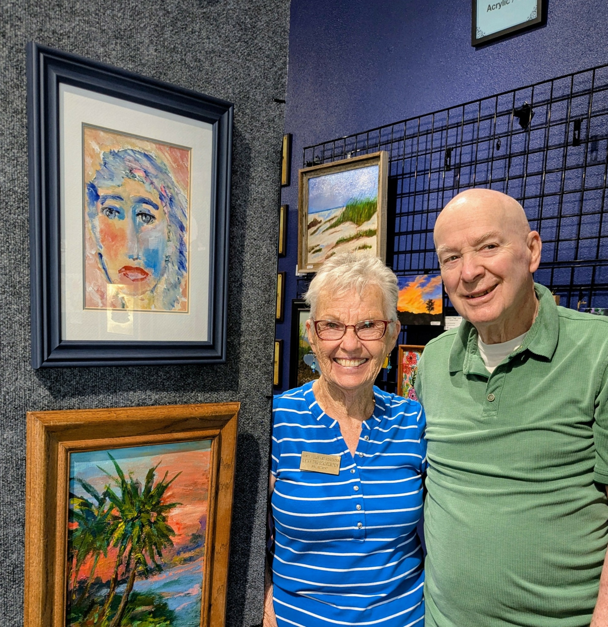 Two people smiling in an art gallery, with framed paintings on the wall behind them.