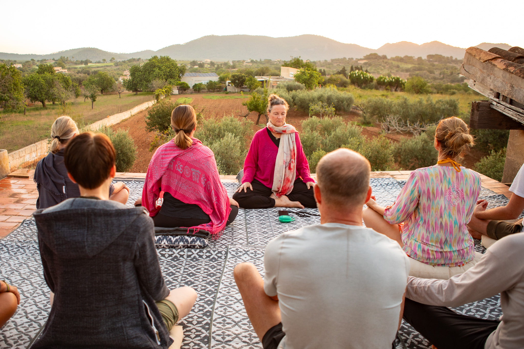 Group of people sitting outdoors on a patterned blanket, facing a woman in a pink jacket, with a scenic landscape background.
