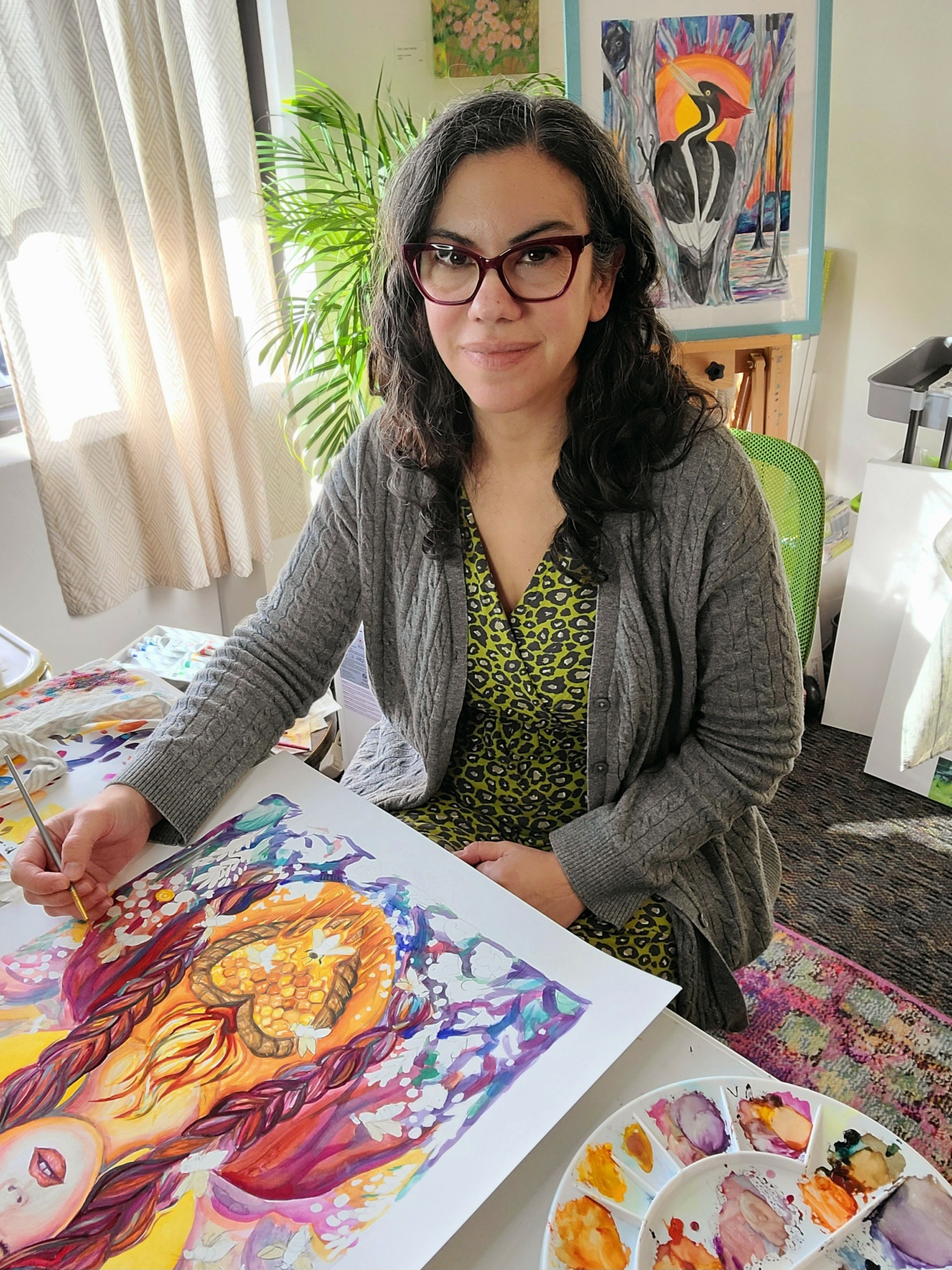 Woman with glasses sitting at a table with colorful artwork and paint palette, smiling at camera.