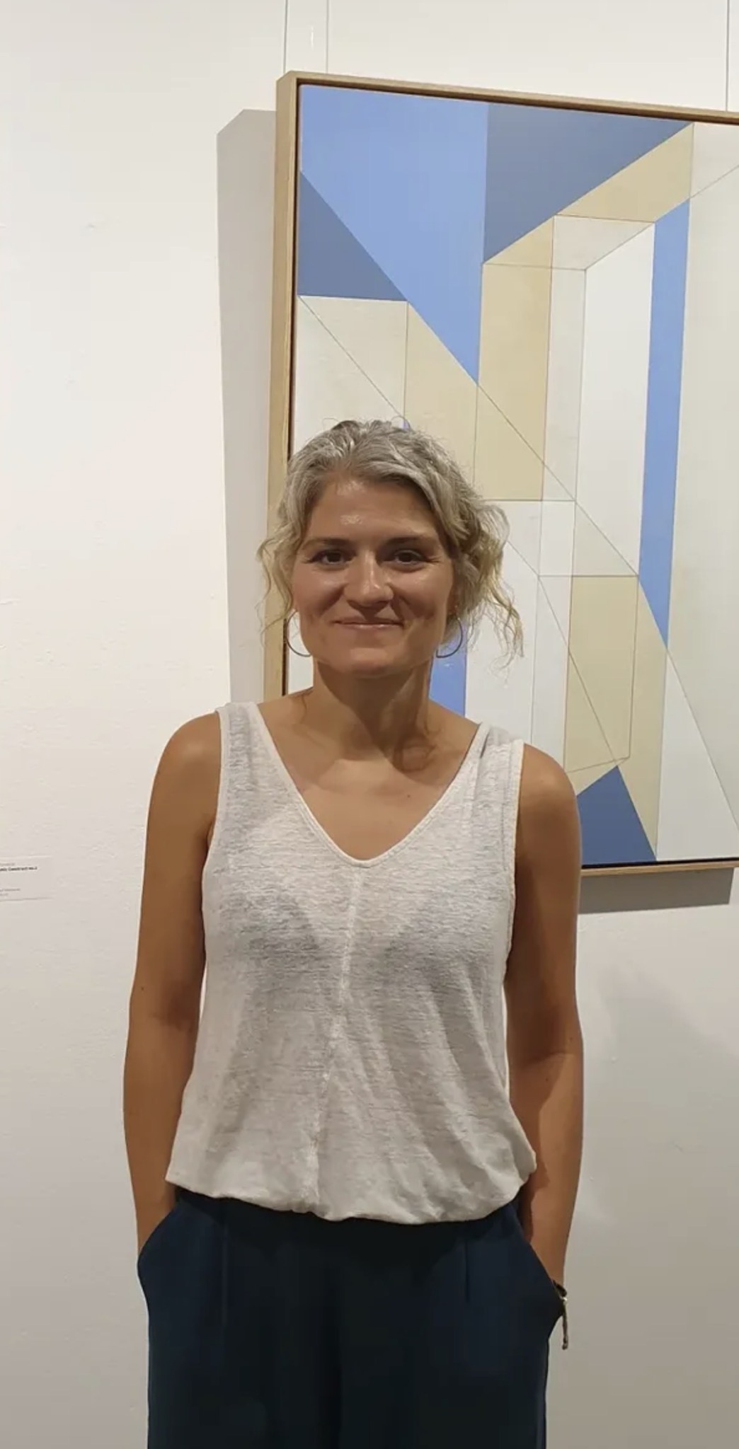 Woman with curly gray hair wearing a sleeveless white top and dark pants, standing in front of an abstract painting.