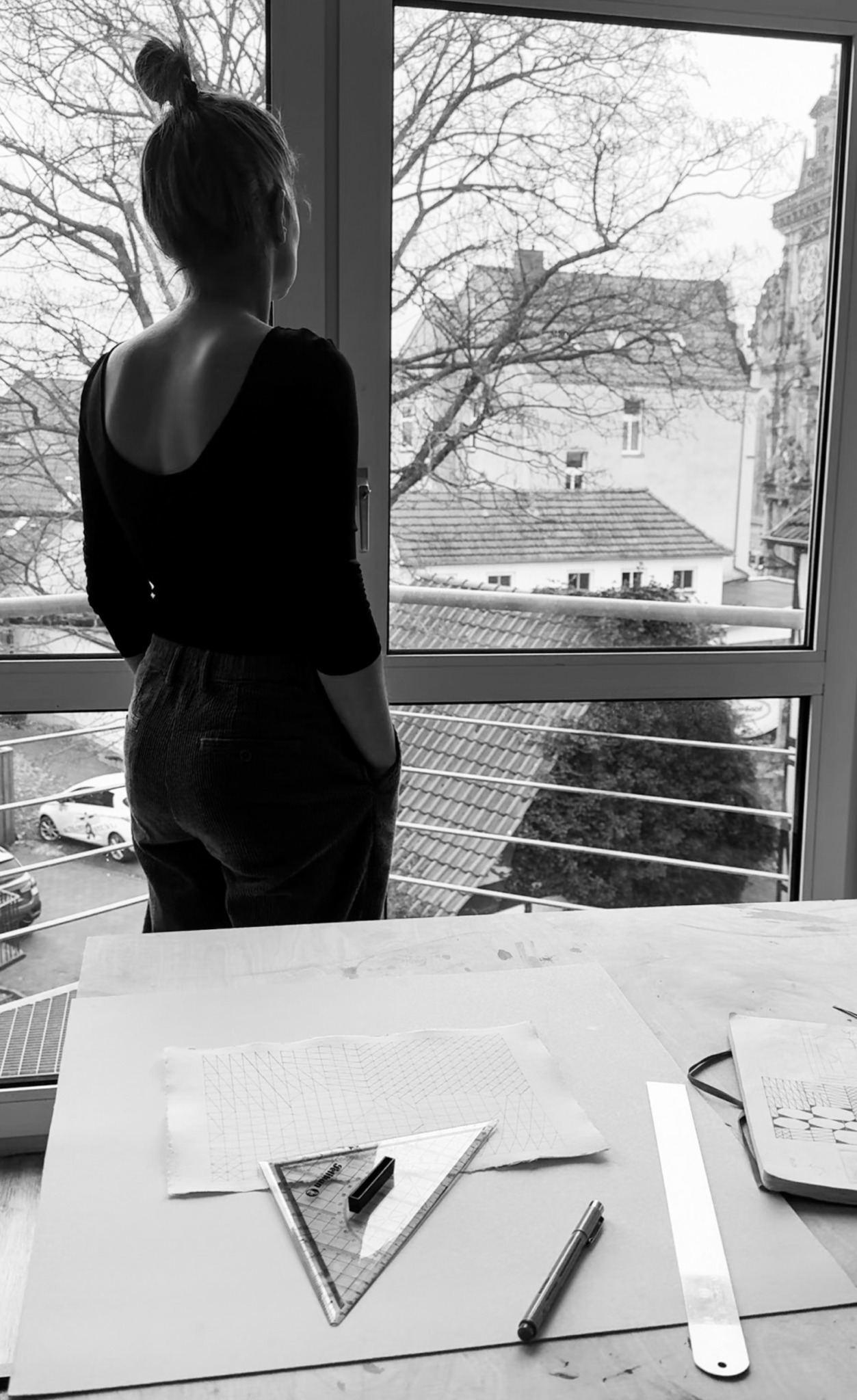 Person standing by window looking outside, with table and stationery in foreground.