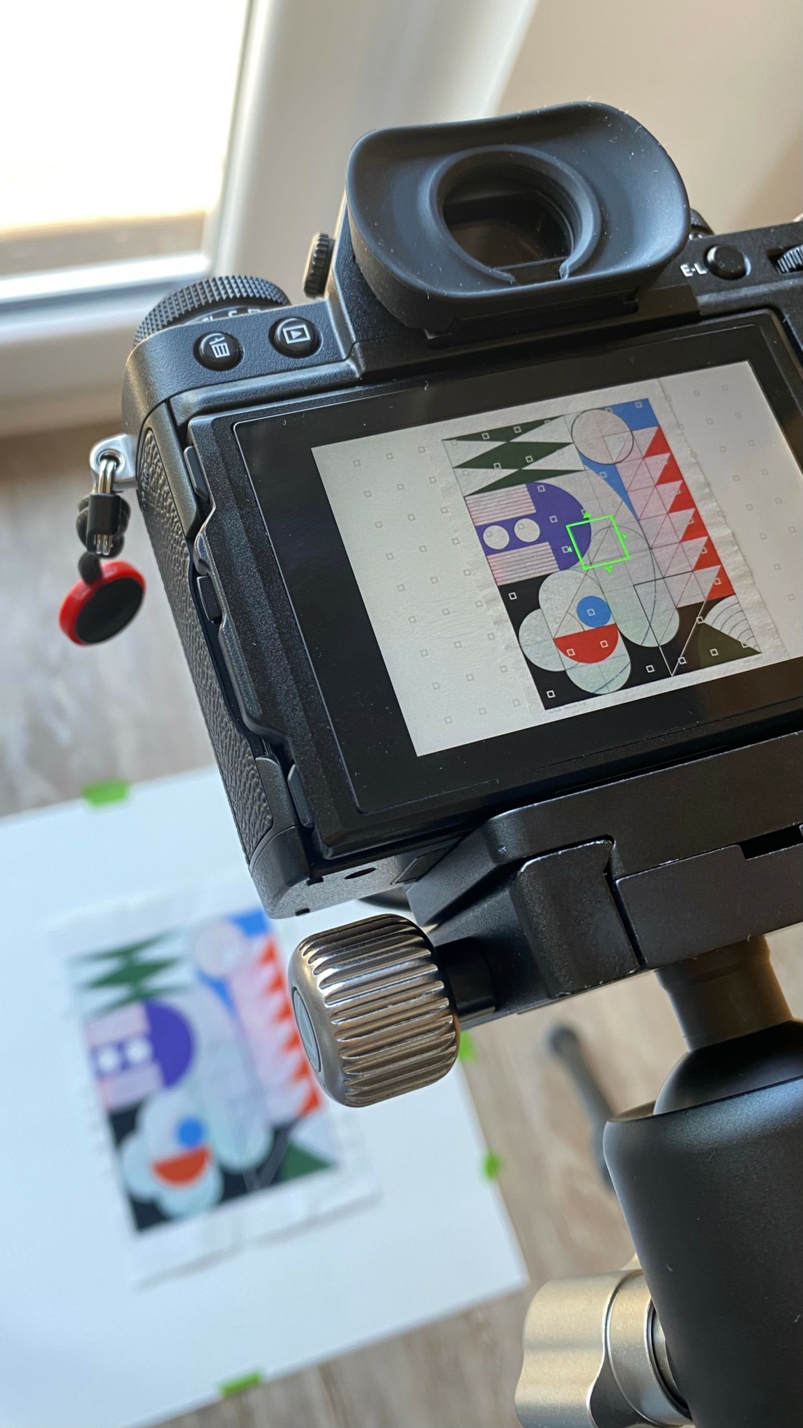 Camera showing a colorful abstract artwork on a screen, with a printed version in the background on a table.