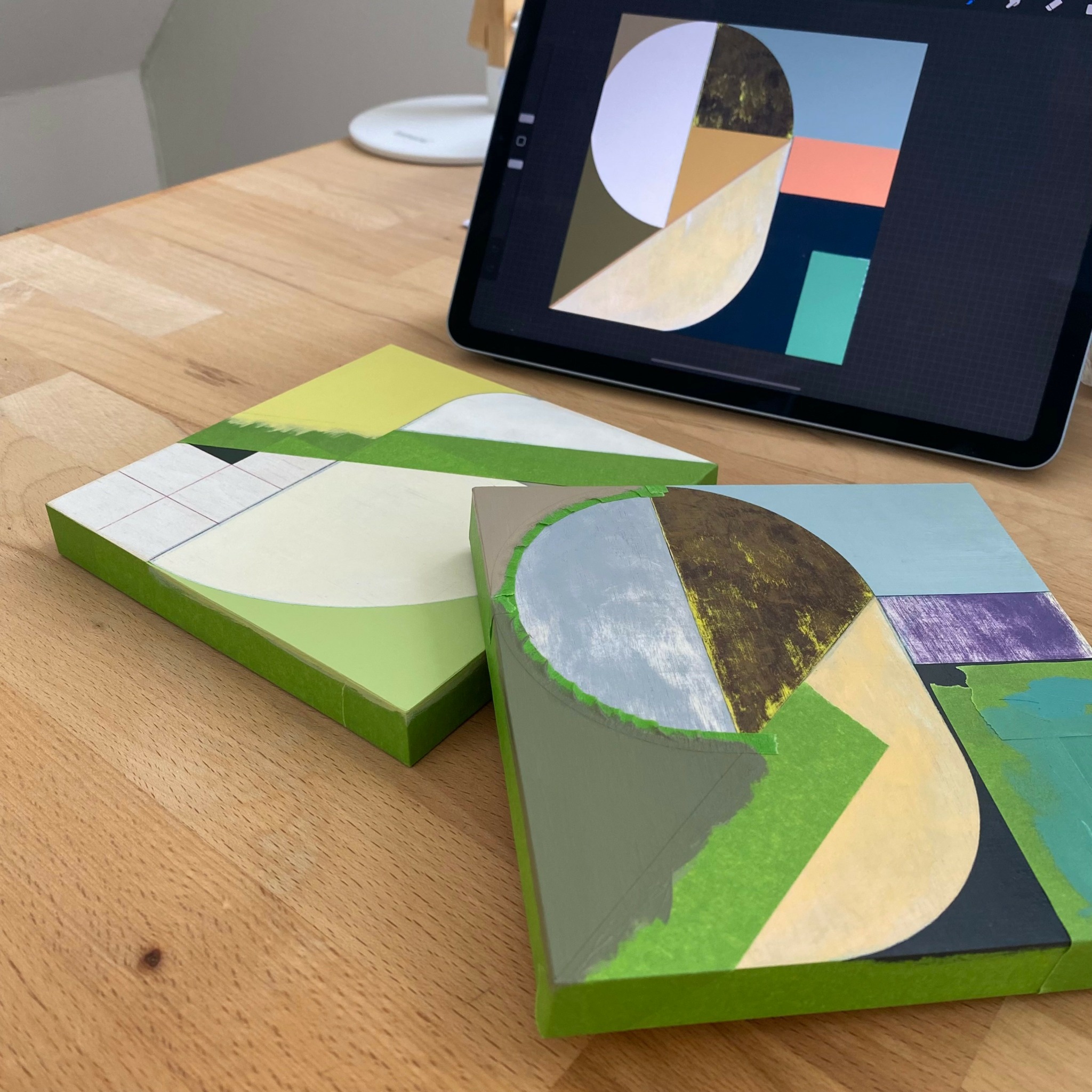 Two abstract landscape paintings on a wooden table with a tablet displaying similar artwork.