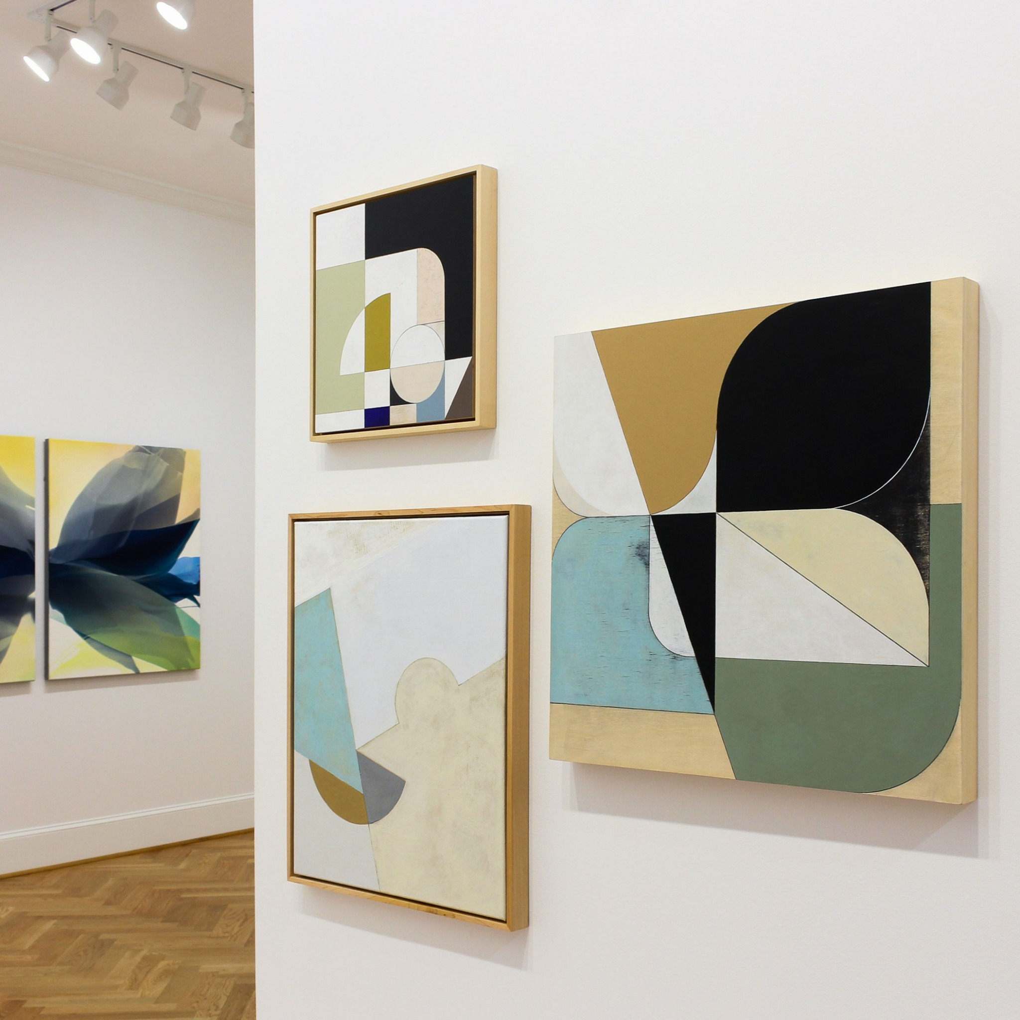 Three abstract paintings on a white wall, with a wooden floor below and ceiling lights above.