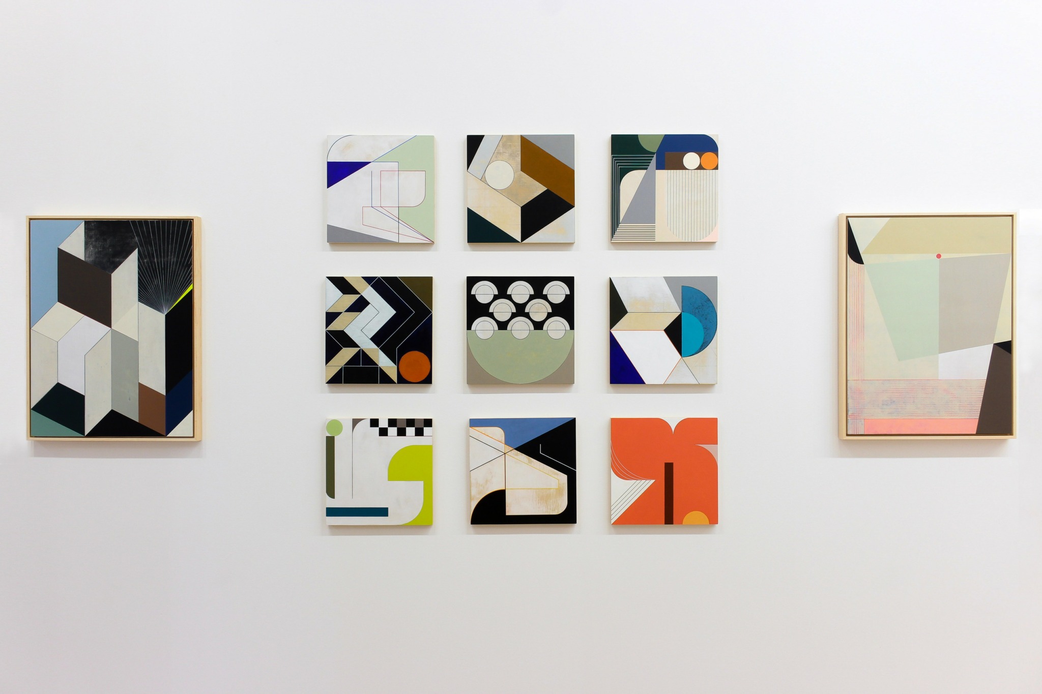 Nine abstract geometric paintings arranged in a grid on a white wall, with two larger paintings on each side.