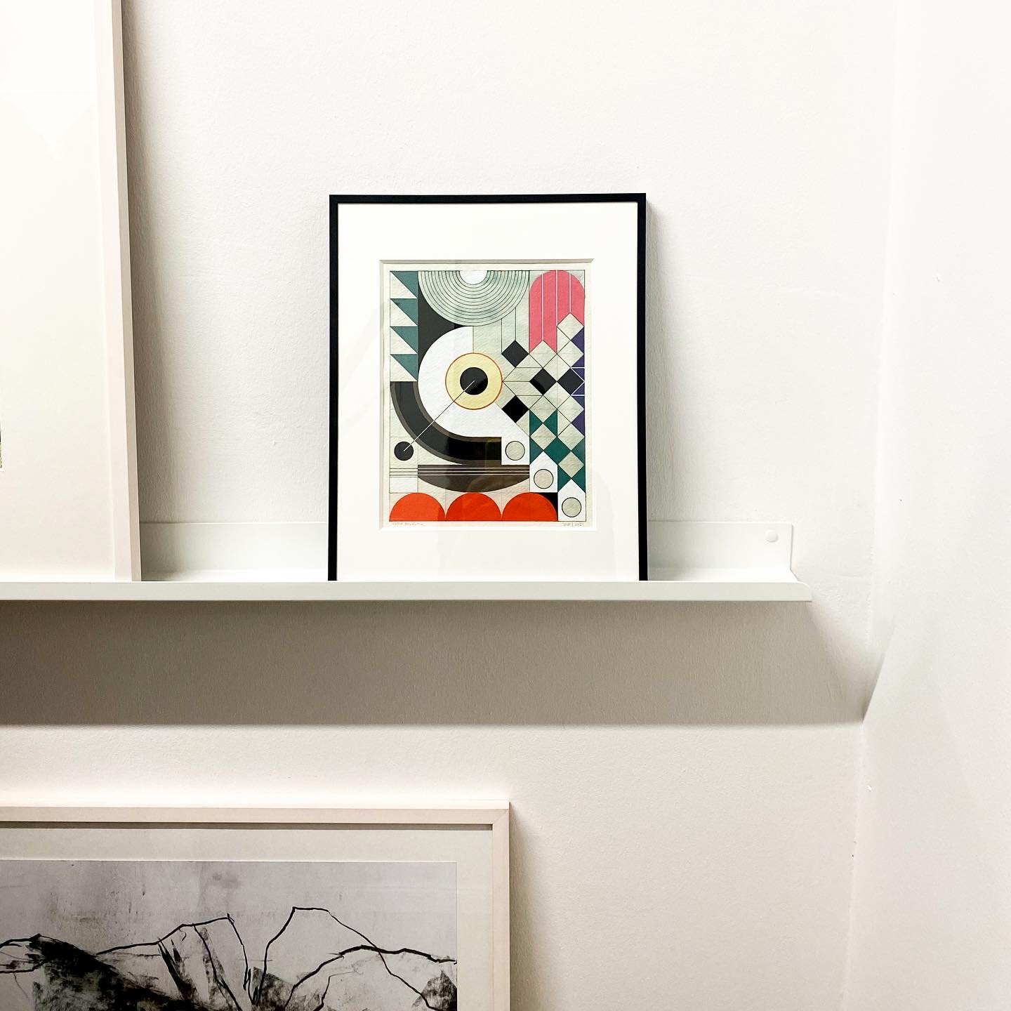 Framed abstract artwork on a white shelf against a white wall.