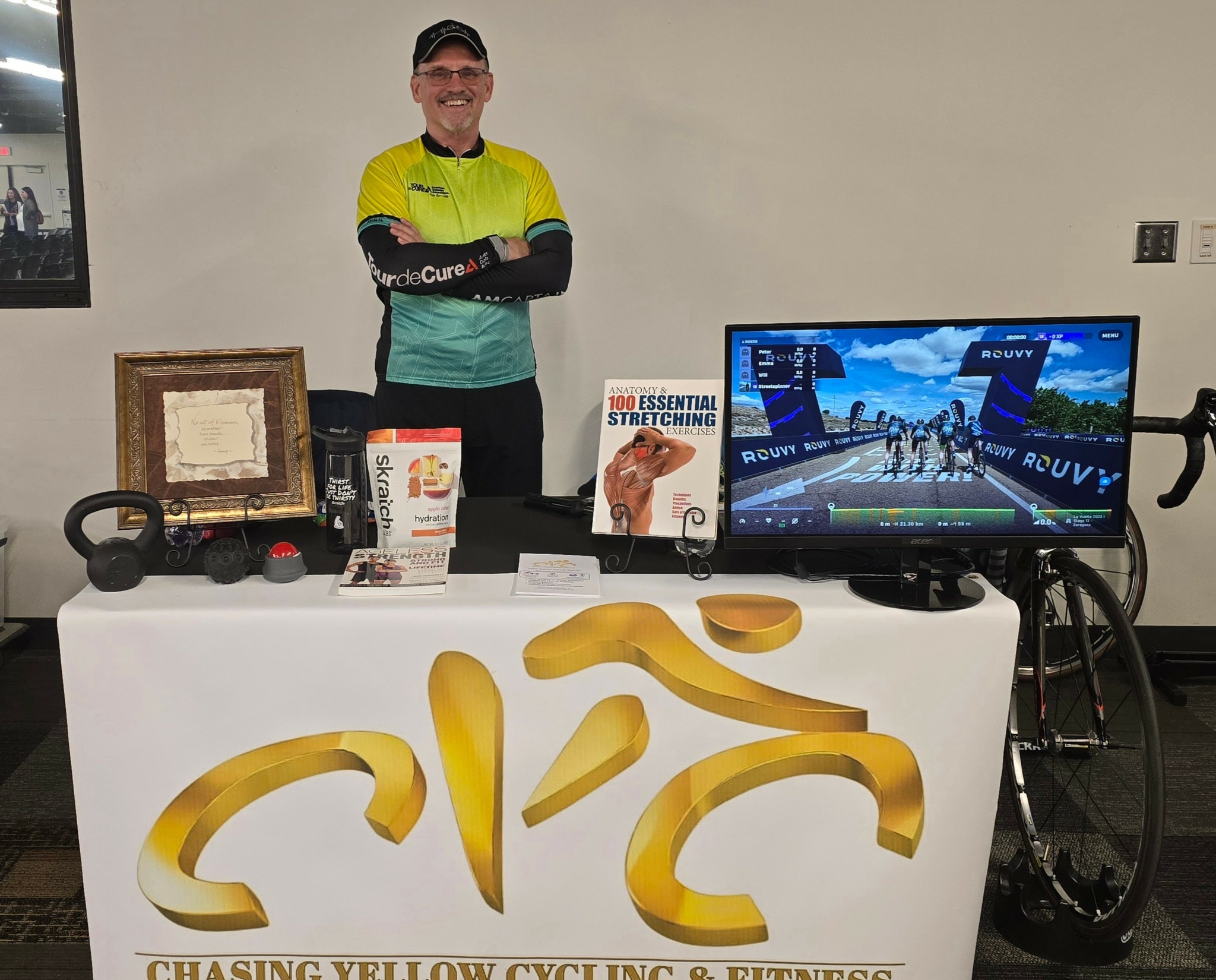 Person standing behind a table with cycling and fitness items, a monitor, and a bicycle wheel in a room.