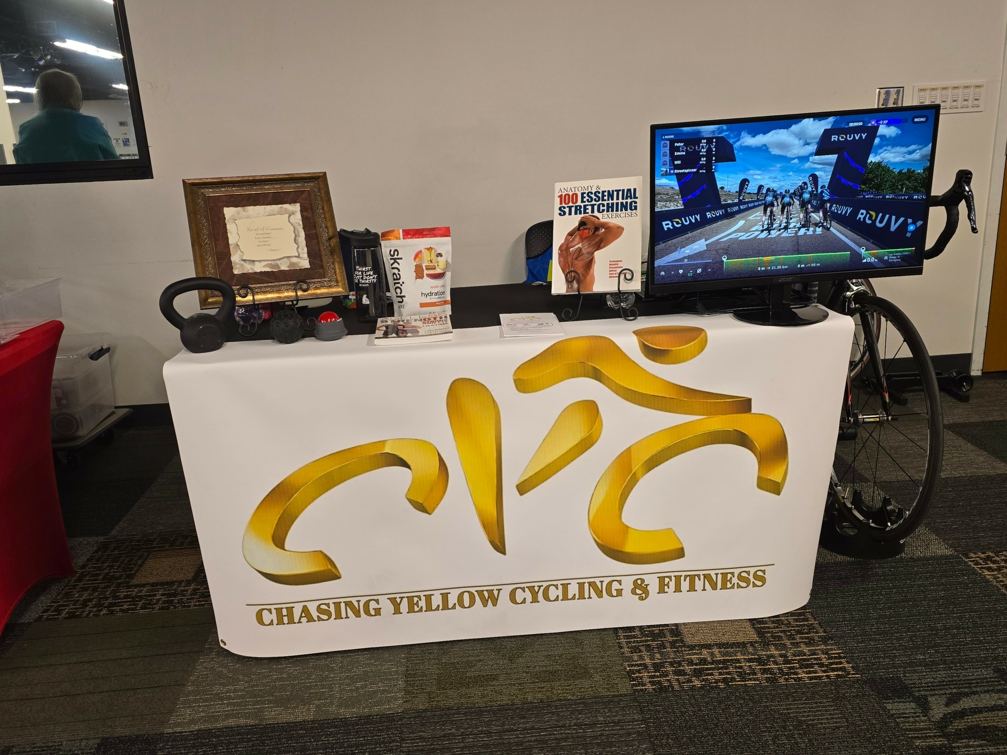 Table with cycling and fitness promotional materials, a monitor, and a framed picture in a room.