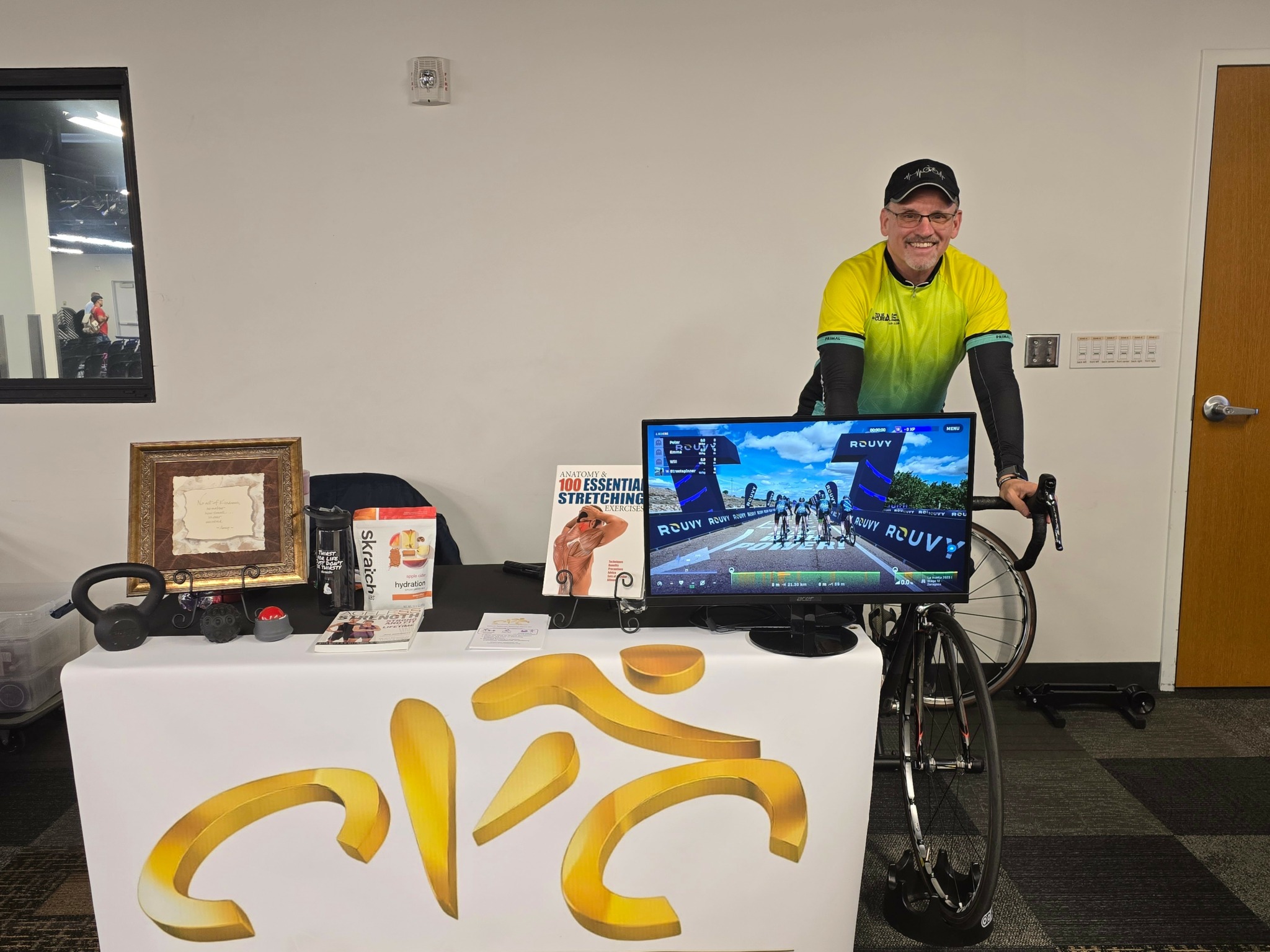 Person in yellow and black cycling jersey standing behind a table with a bicycle, monitor, and items in a room.