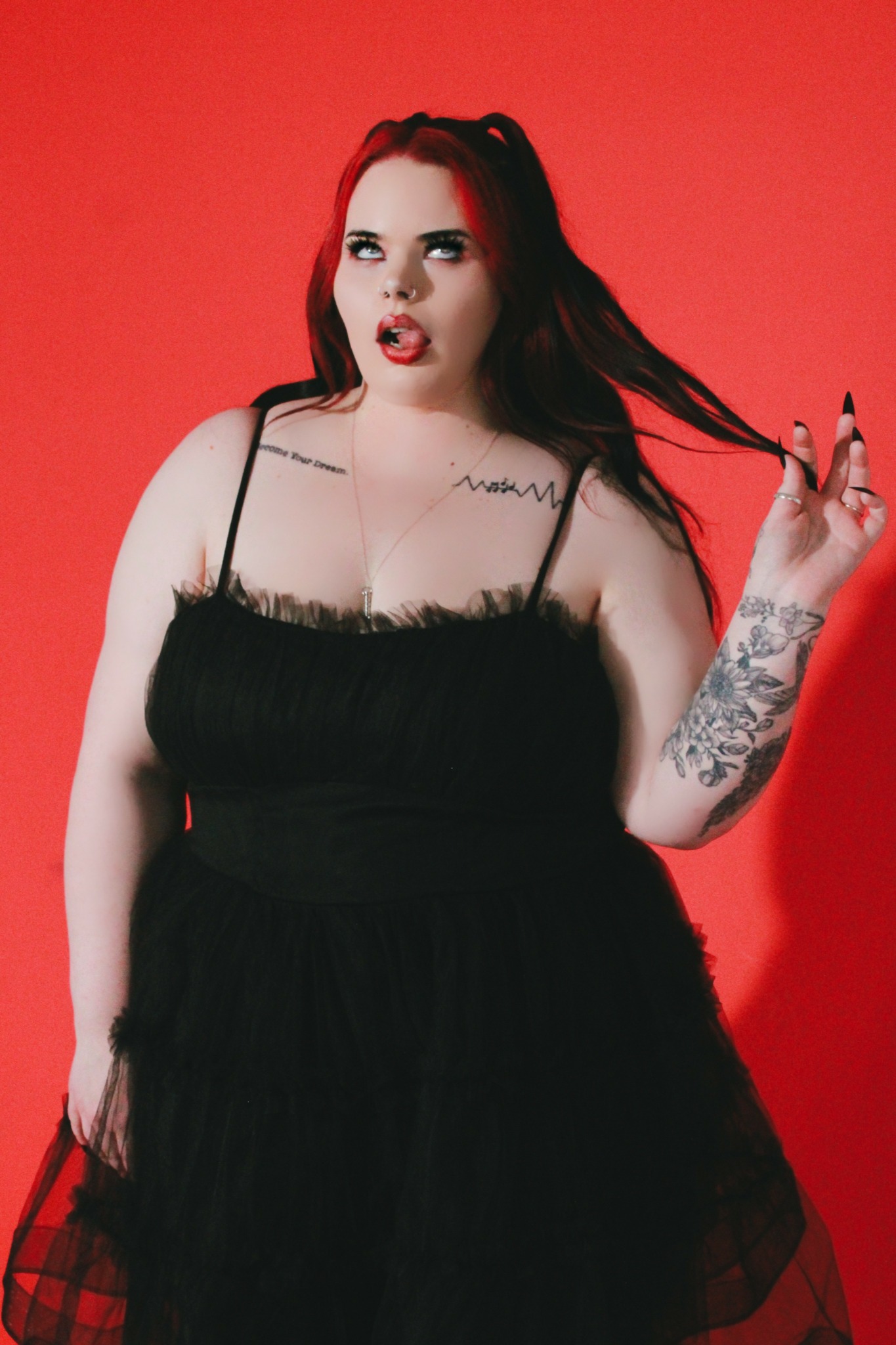 Woman with long dark hair, tattoos, wearing a black dress, standing against red background.