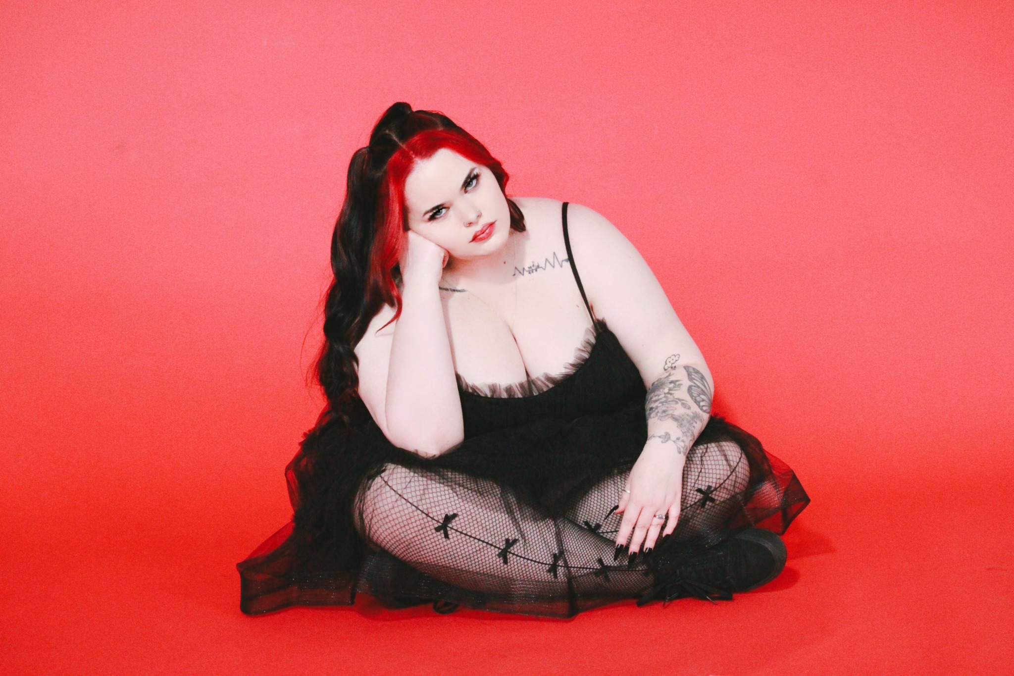 Woman with long dark hair, tattoos, and makeup, sitting on a red background, wearing a black dress and fishnet stockings.