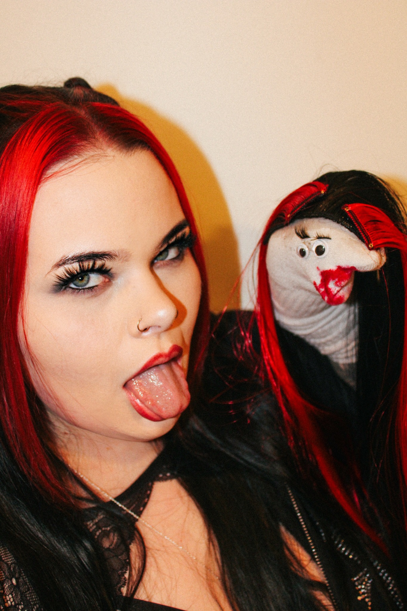Woman with red hair sticking out her tongue, wearing makeup and a nose ring, next to a doll with red and black hair.