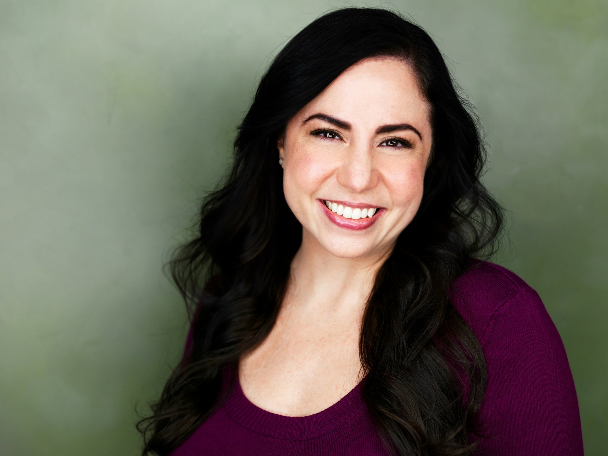 Woman with long dark hair smiling, wearing a purple top, against a green background.