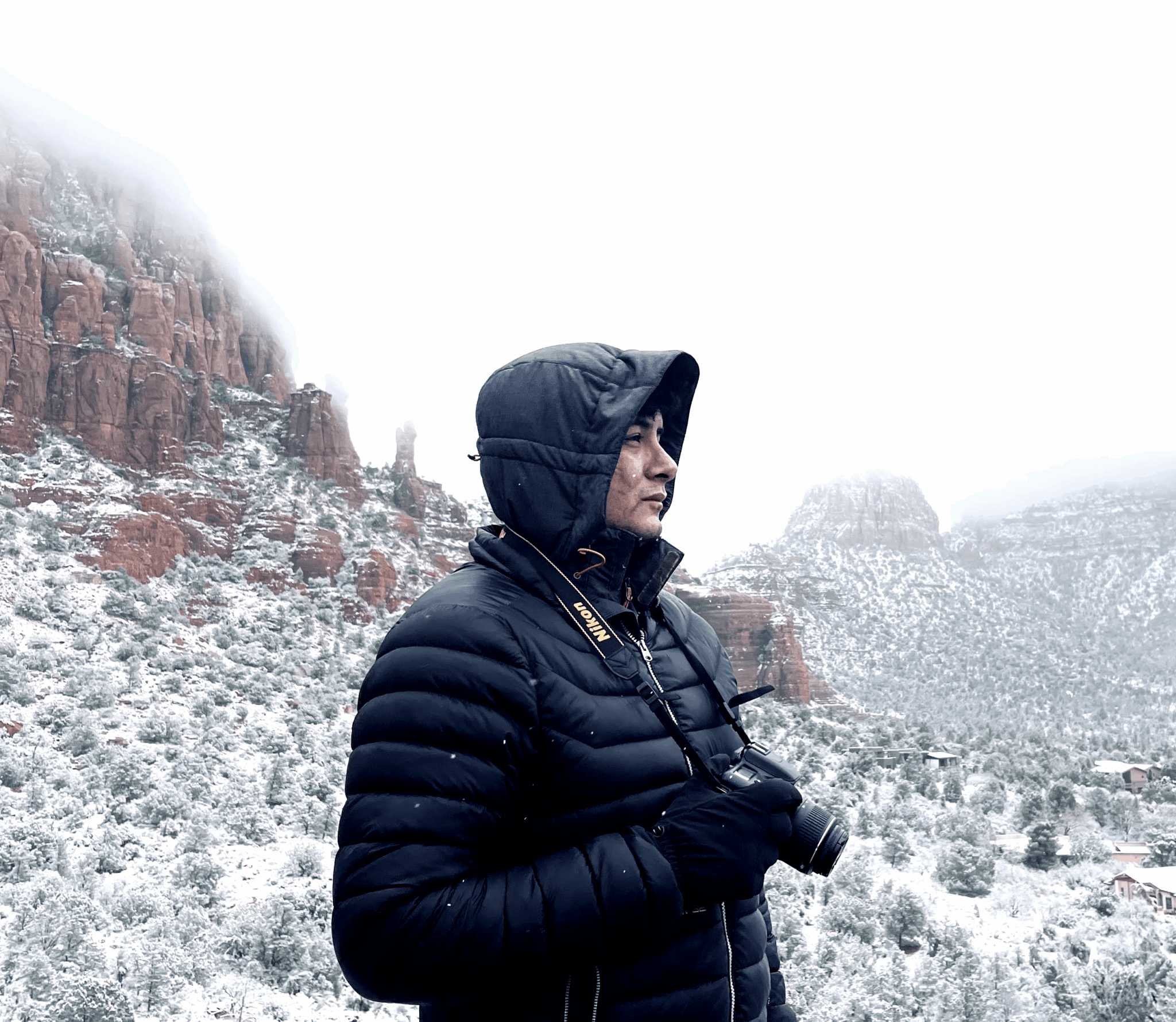 Person in black jacket and hood holding a camera, snowy mountain landscape background.