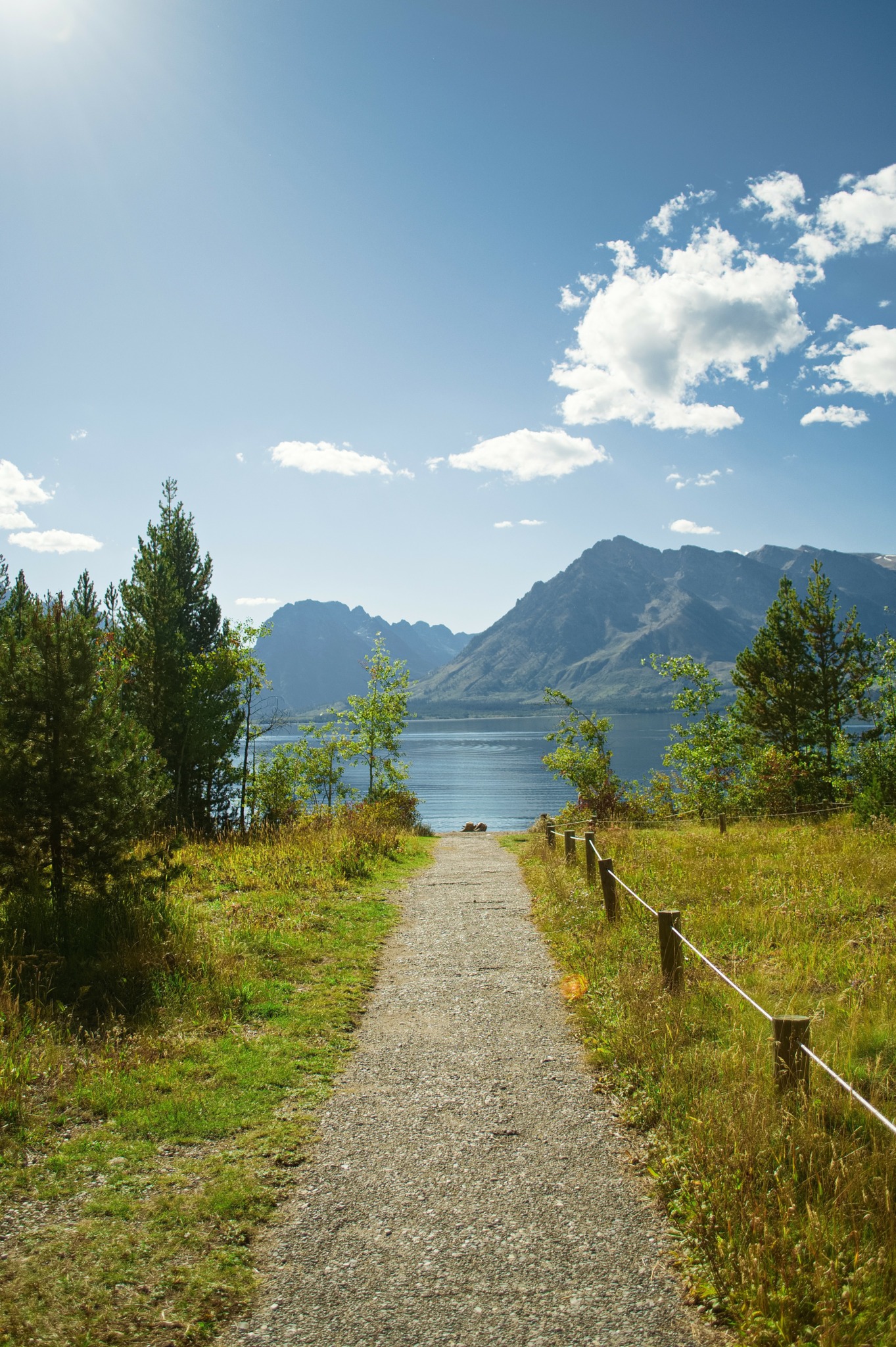 Gravel path leading to a lake with mountains and trees under a partly cloudy sky.