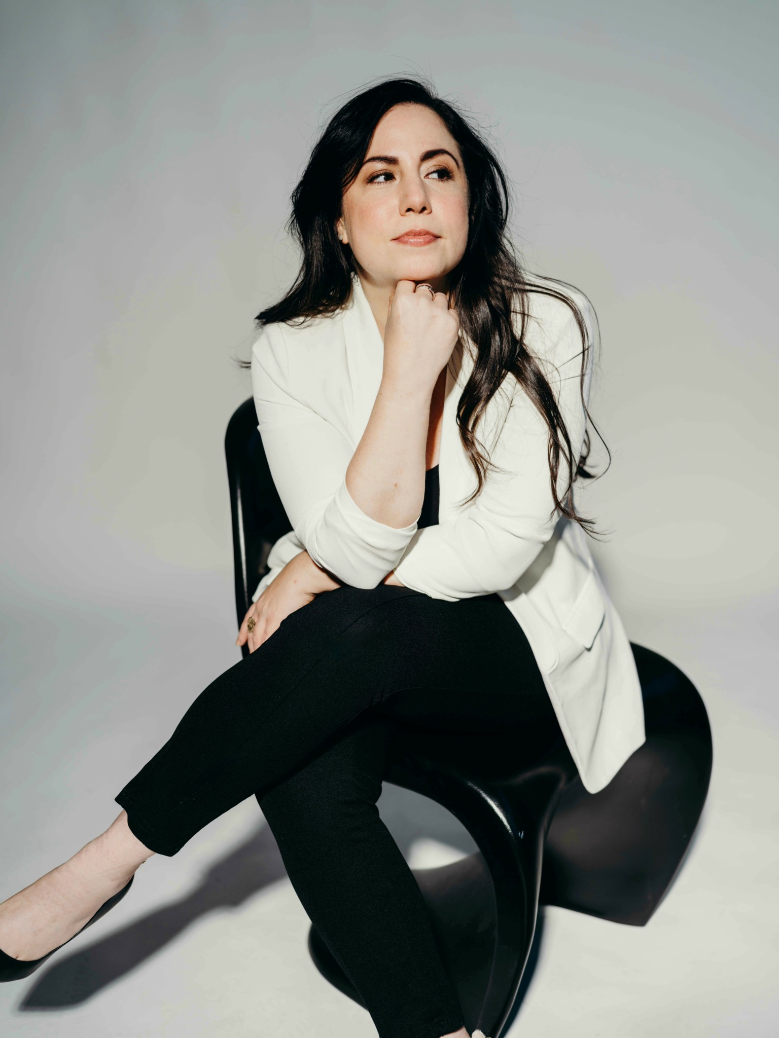 Woman with long dark hair sitting on a black chair, wearing a white blazer and black pants, looking thoughtful.