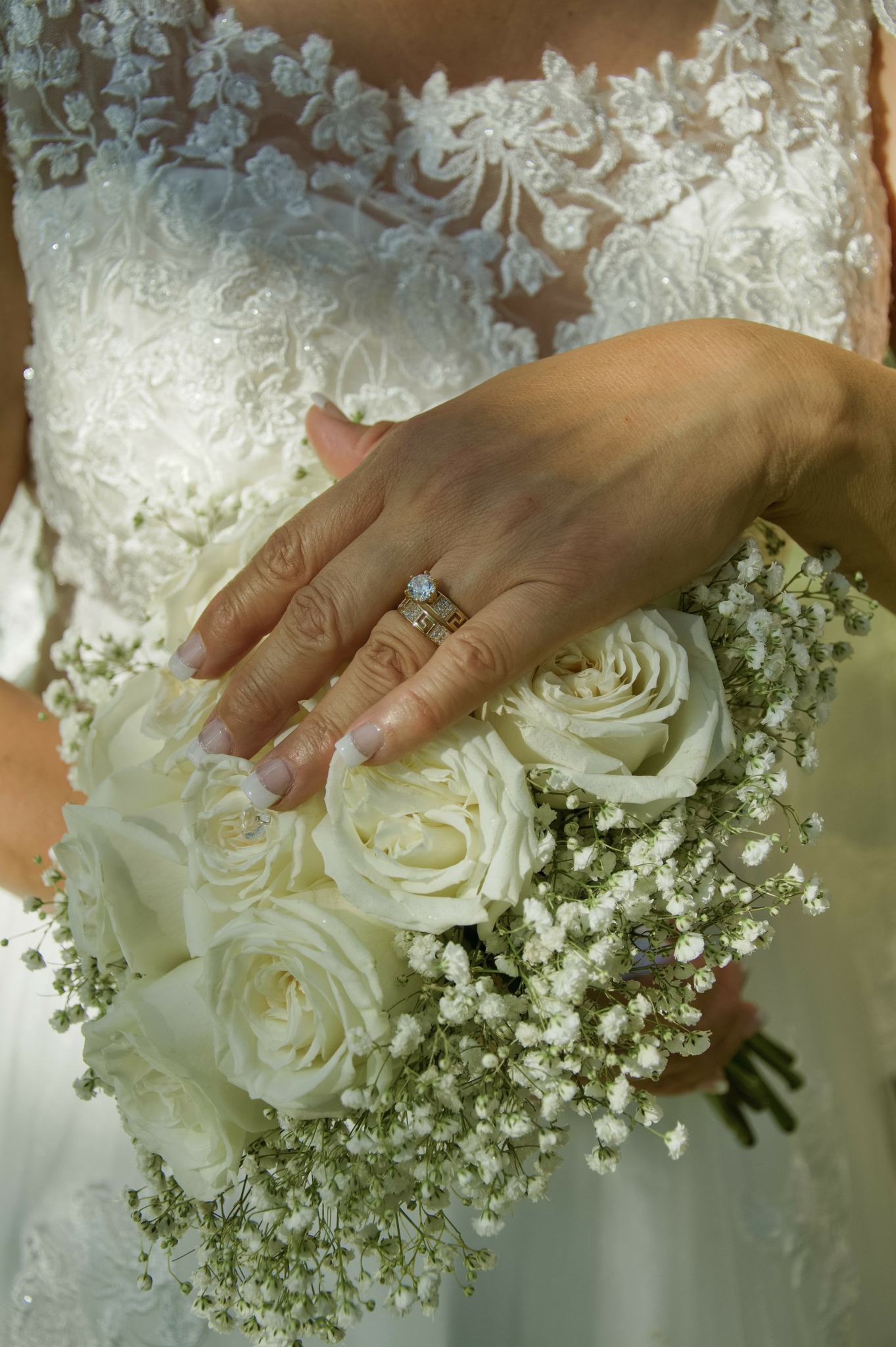Hand with wedding ring resting on bouquet of white roses and baby's breath, bride in lace dress in background.