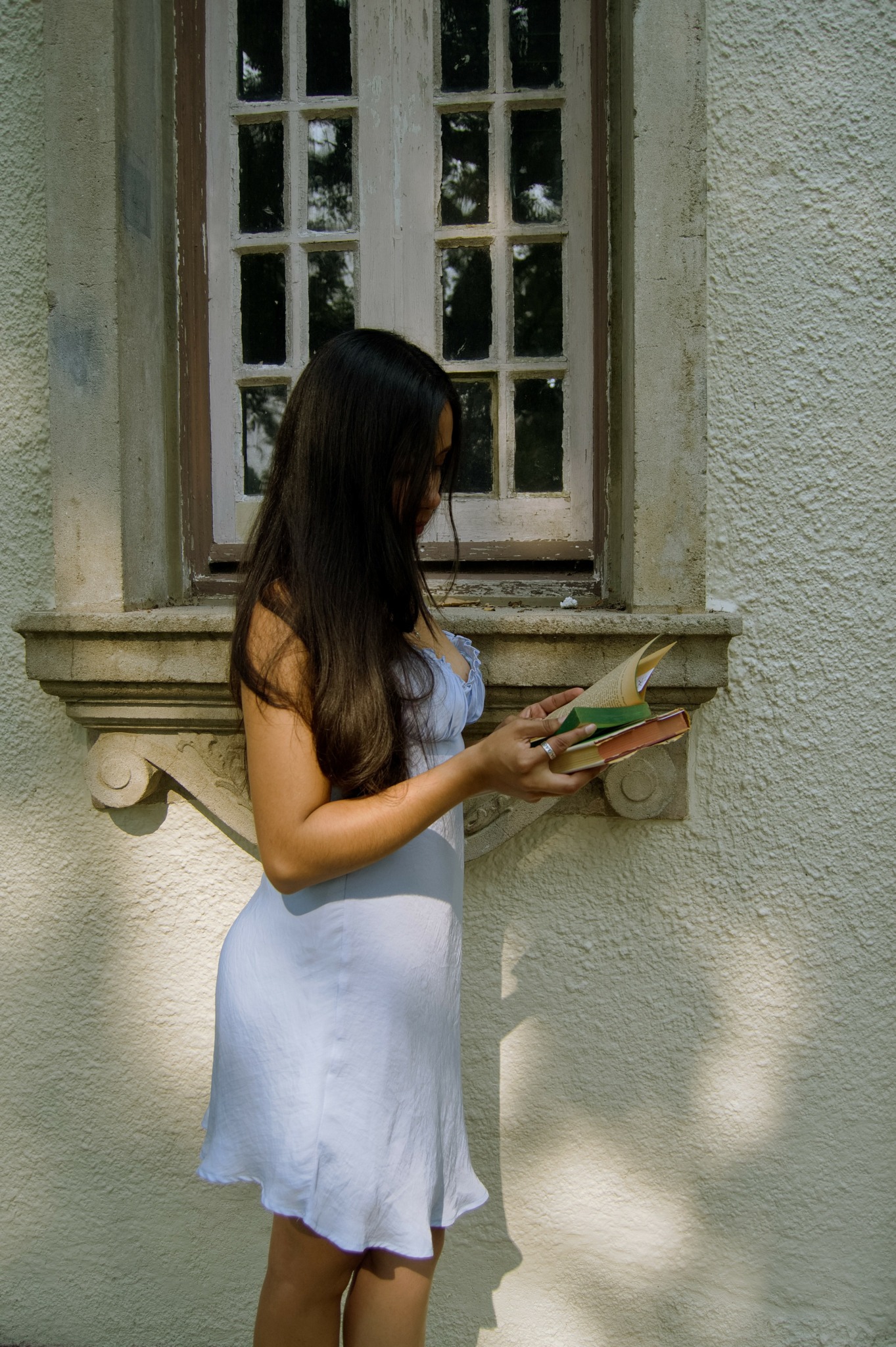 Woman with long dark hair reading a book outside near a window on a textured wall.