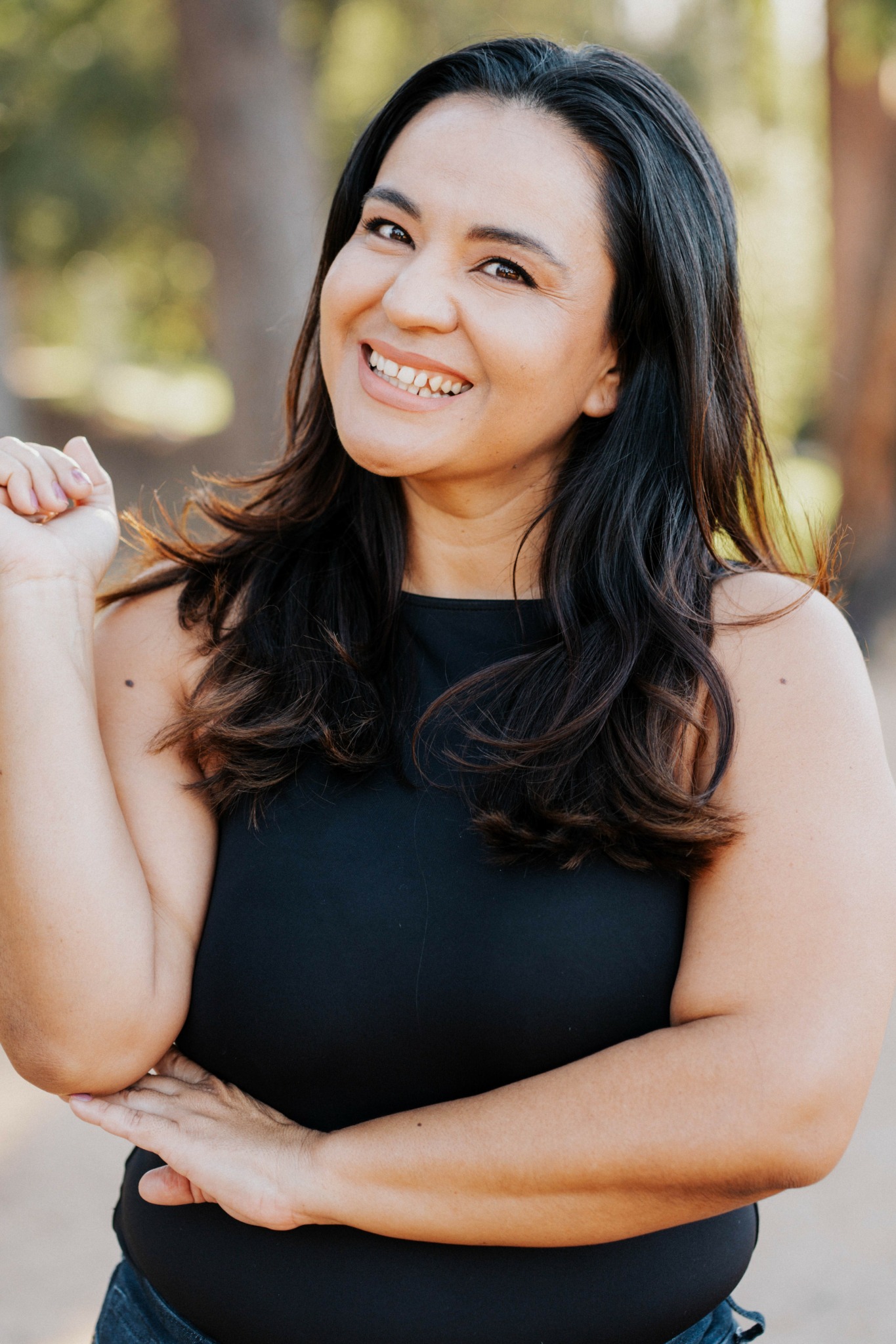 Smiling woman with long dark hair outdoors, wearing a black sleeveless top, in natural light.