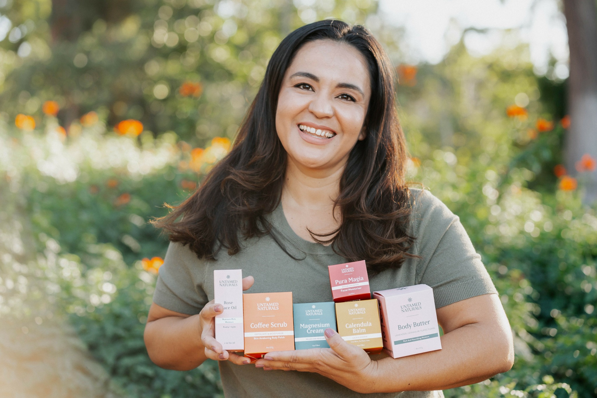 Woman holding colorful boxes outdoors, smiling, with trees and flowers in background.