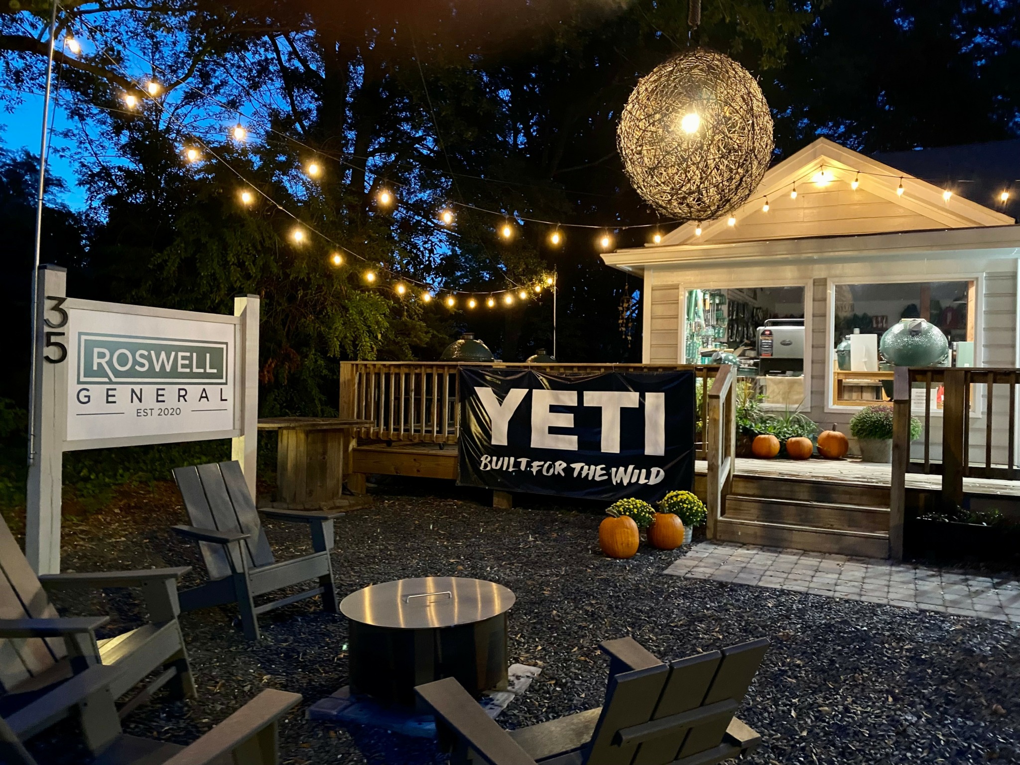Outdoor area with string lights, a large spherical decoration, pumpkins, and a small building with a person inside.