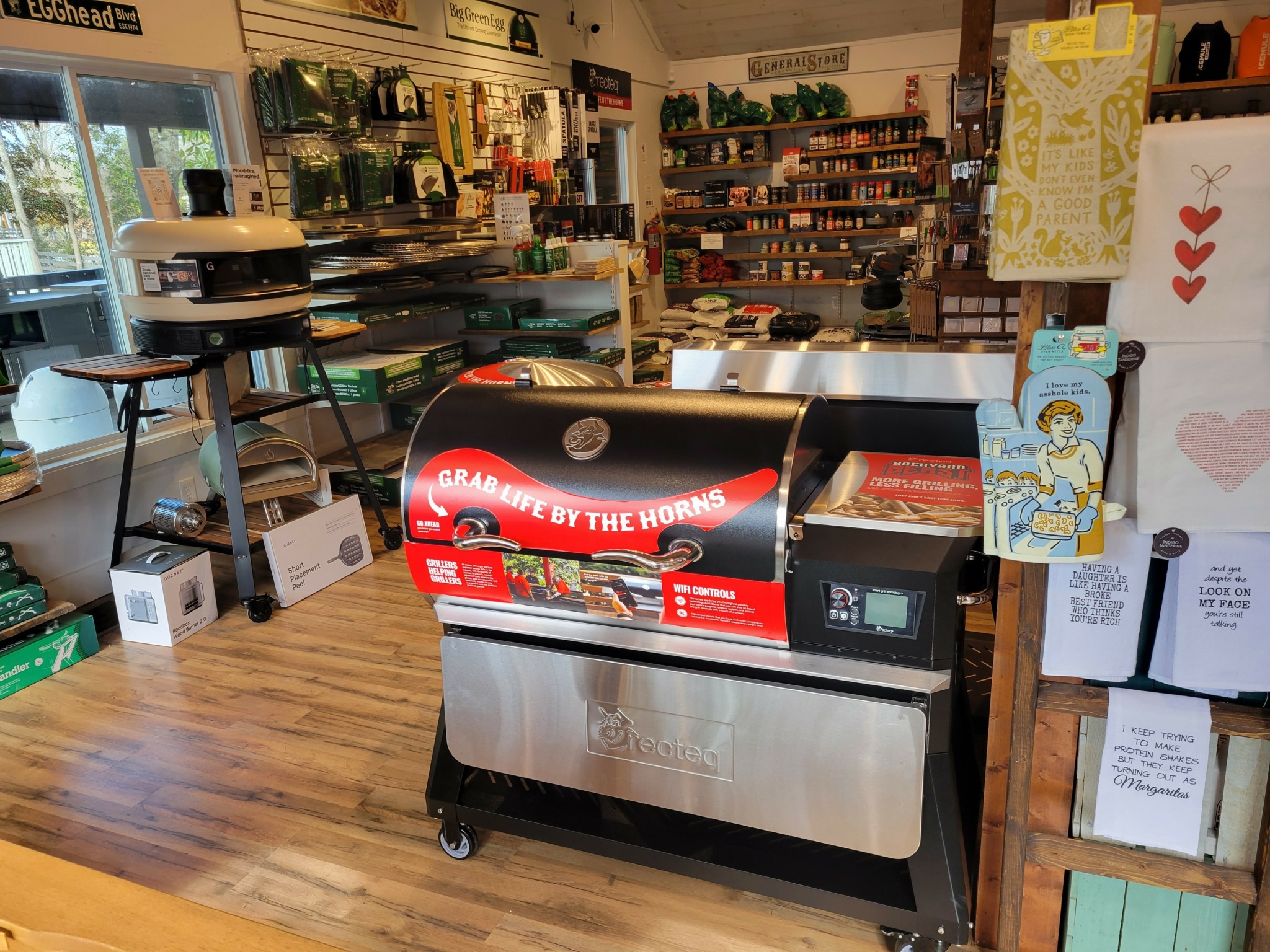 Indoor store with shelves of various products, a large black and red grill in the center, and wooden flooring.