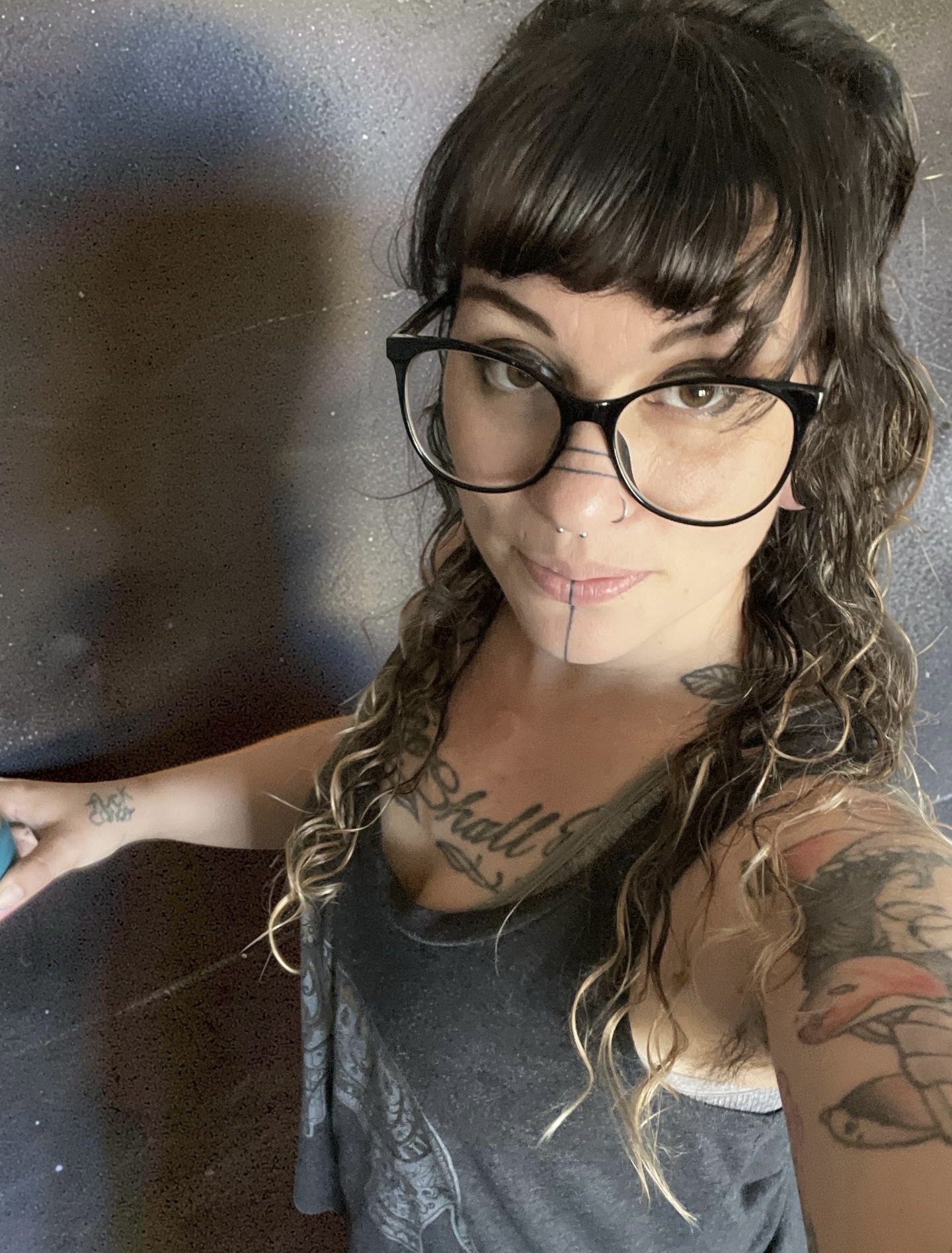 Woman with glasses and tattoos, curly hair, smiling, wearing a dark top, standing against a dark background.