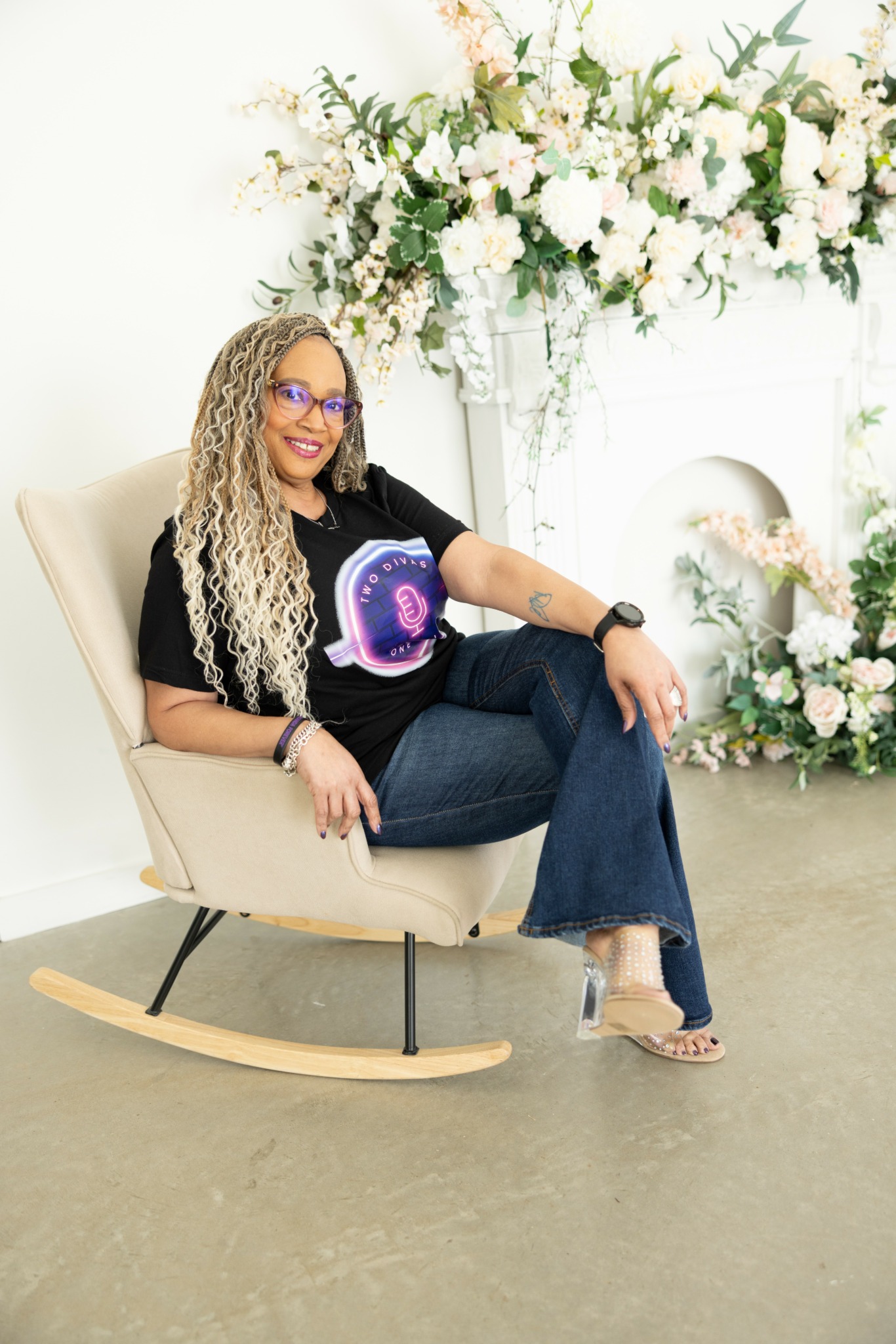 Woman with curly hair and glasses sitting on a beige rocking chair, smiling, with floral arrangements behind her.