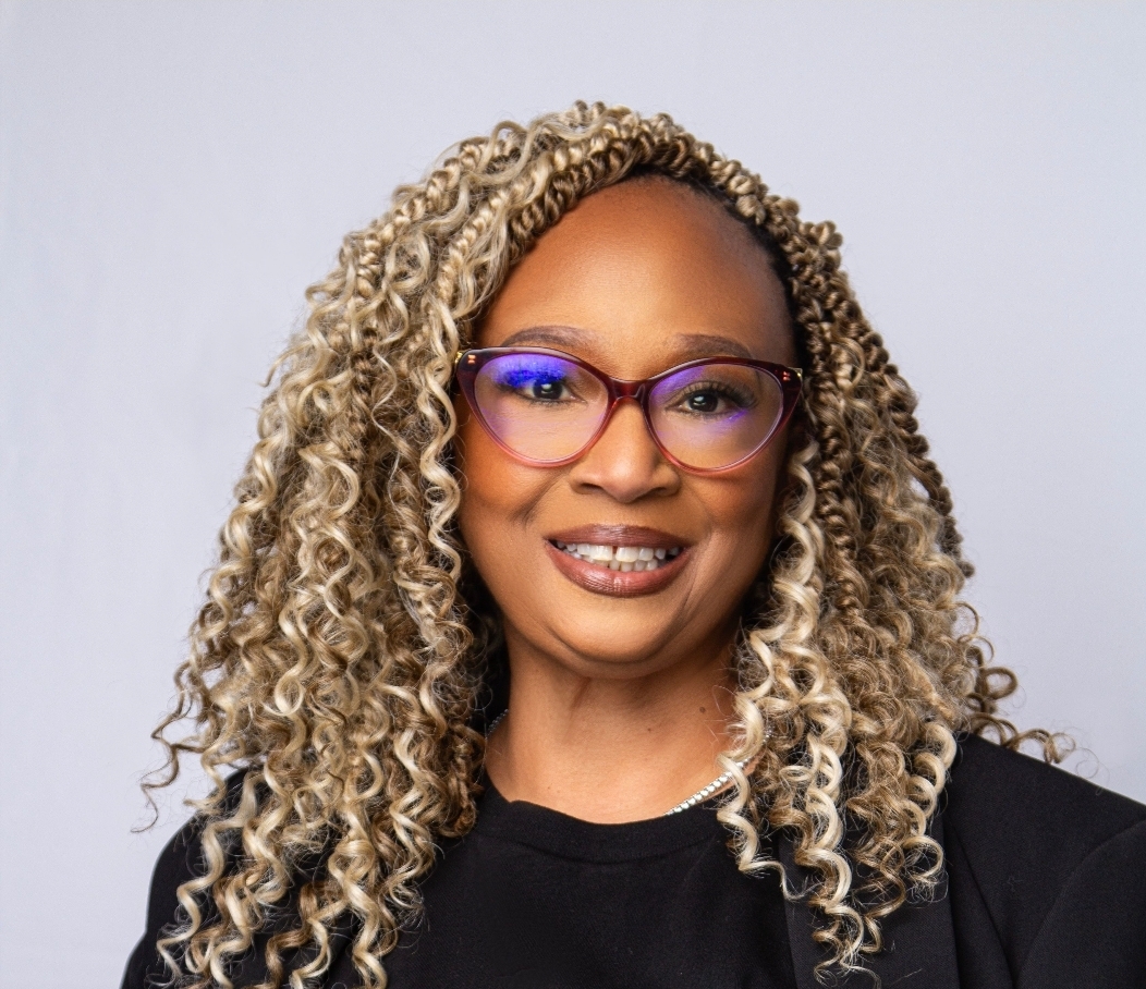 Woman with curly blonde hair, glasses, smiling, wearing a black top, against a plain background.