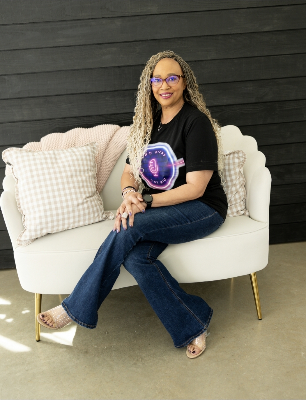 Woman with curly hair and glasses sitting on a white sofa with checkered pillows, smiling, black wall background.