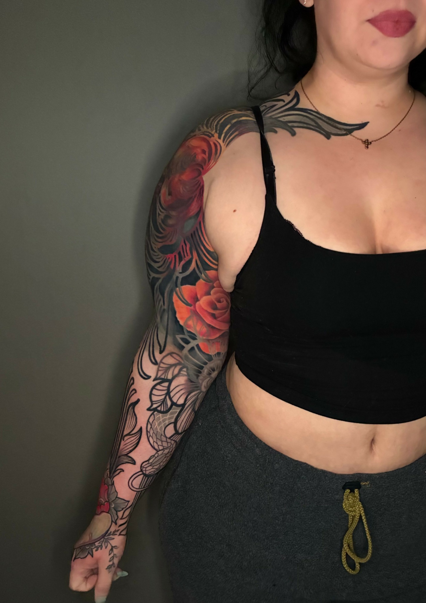 Woman with black hair, wearing a black crop top and gray pants, showing colorful floral tattoos on her arm and shoulder.