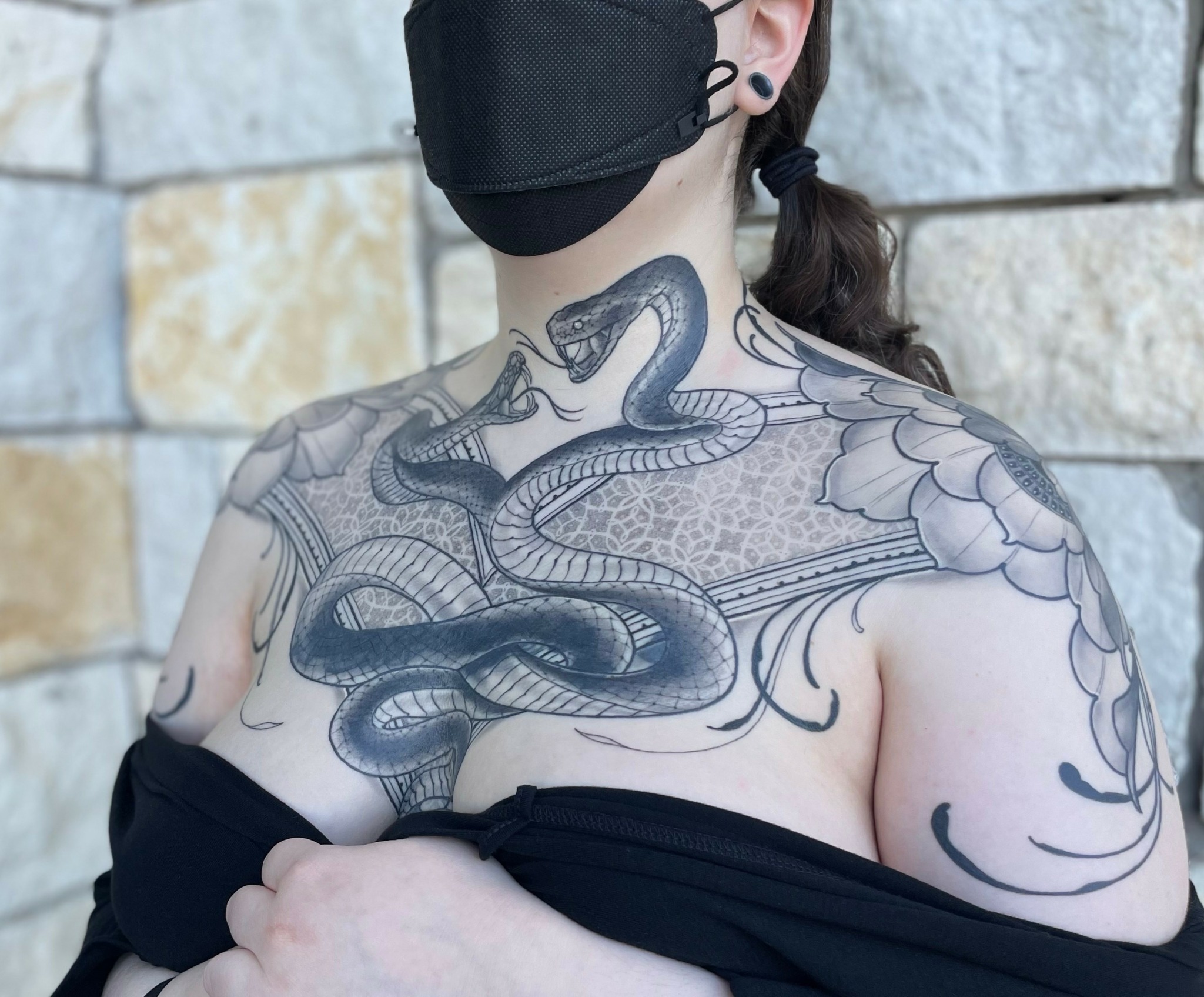 Person with a large snake tattoo on chest and shoulders, wearing a black face mask and black clothing off shoulder.