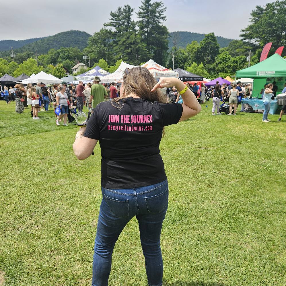 Person with short hair wearing a black T-shirt and jeans at outdoor event with tents and crowd.