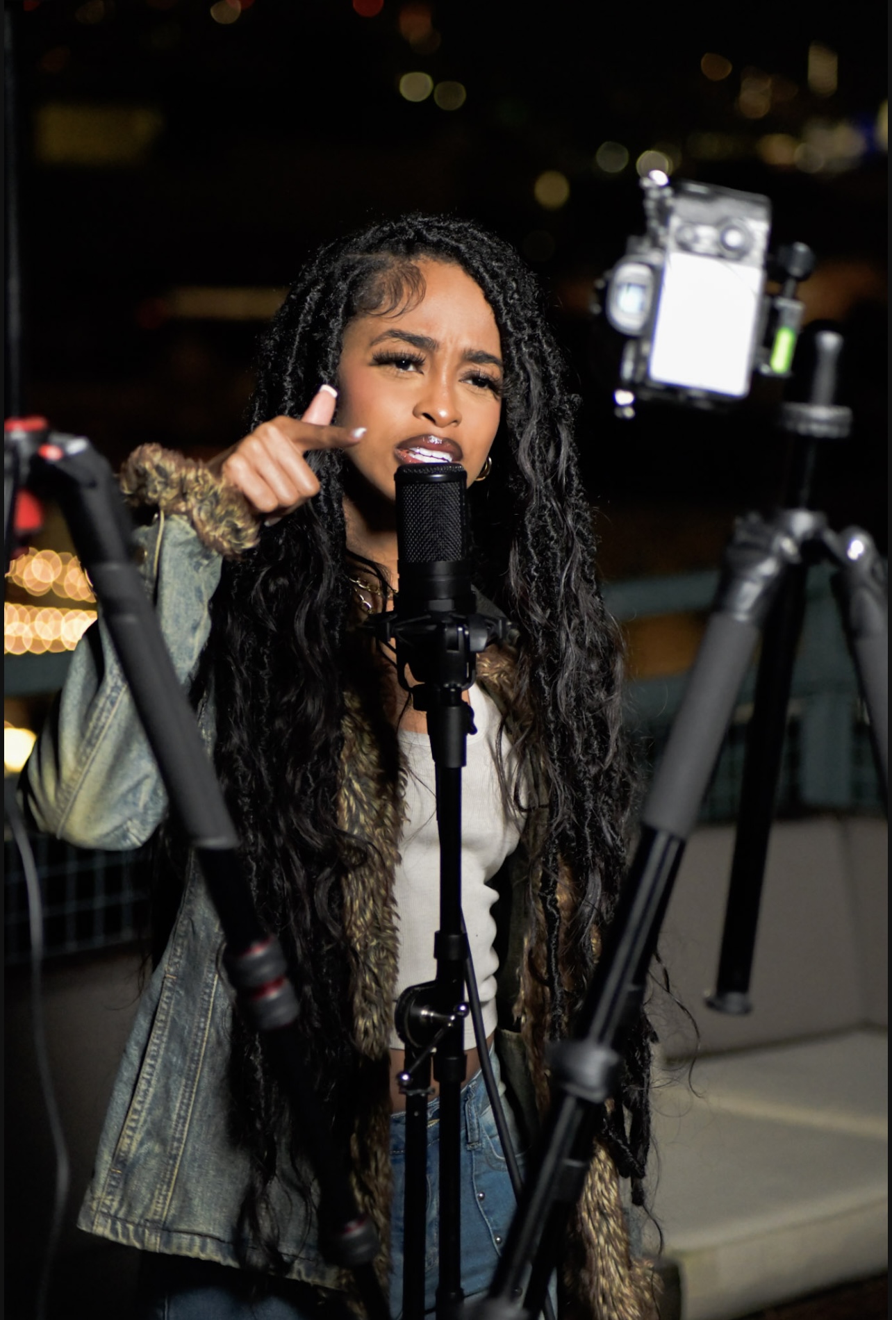 Young woman with long curly hair speaking into a microphone, surrounded by recording equipment outdoors at night.