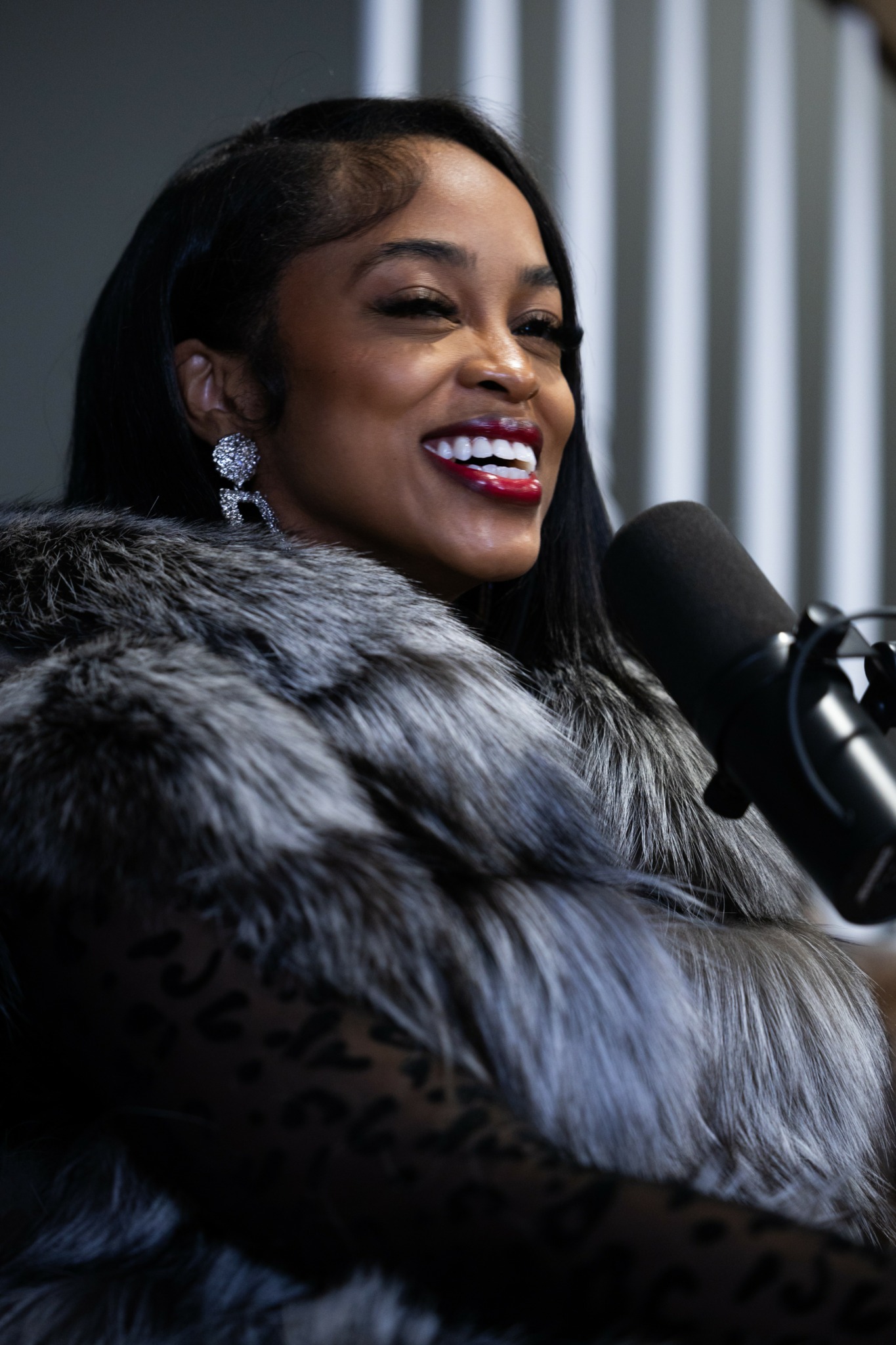 Woman with dark hair and earrings speaking into a microphone, wearing a fur coat, smiling.