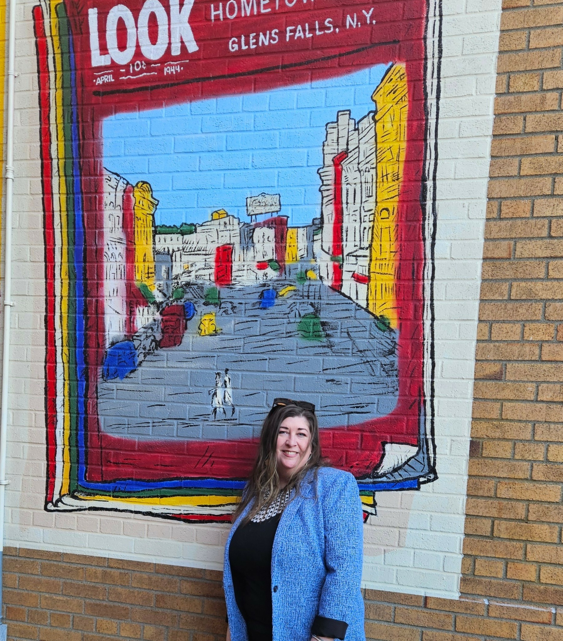 Woman with long hair wearing a blue coat standing in front of a colorful mural of a city street scene on a brick wall.