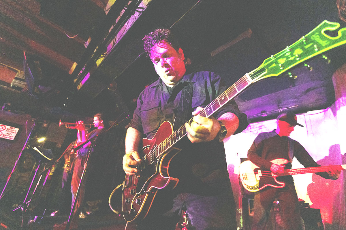 Musician playing an electric guitar on stage with other band members in background.