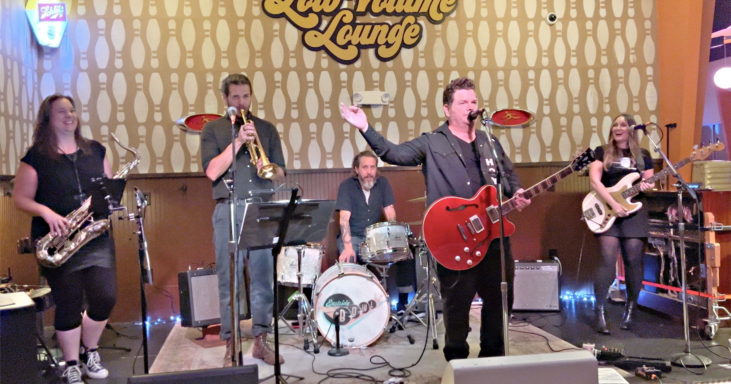 Band performing on stage with five members, musical instruments, and a patterned backdrop.