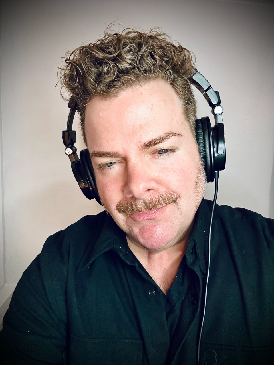 Man with curly hair and mustache wearing headphones and a black shirt, looking down.