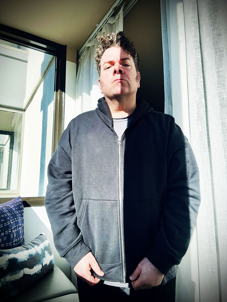 Young man standing indoors near window with sunlight, wearing a dark zip-up hoodie, looking directly at camera.