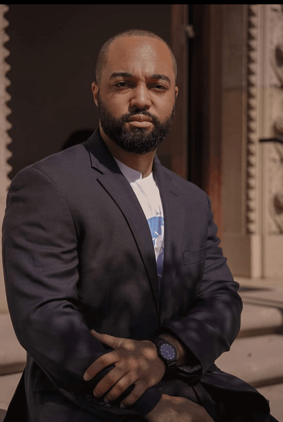 Man with beard in a dark blazer and white shirt, standing outdoors with architectural background.